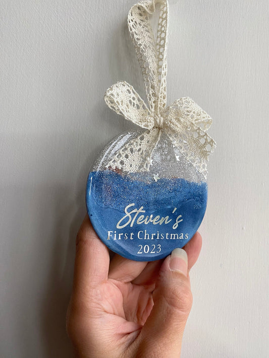 Hand holding a Blue Baby First Christmas resin bauble with glittery silver top, twinkling stars, personalised silver lettering, and beige lace ribbon with bow