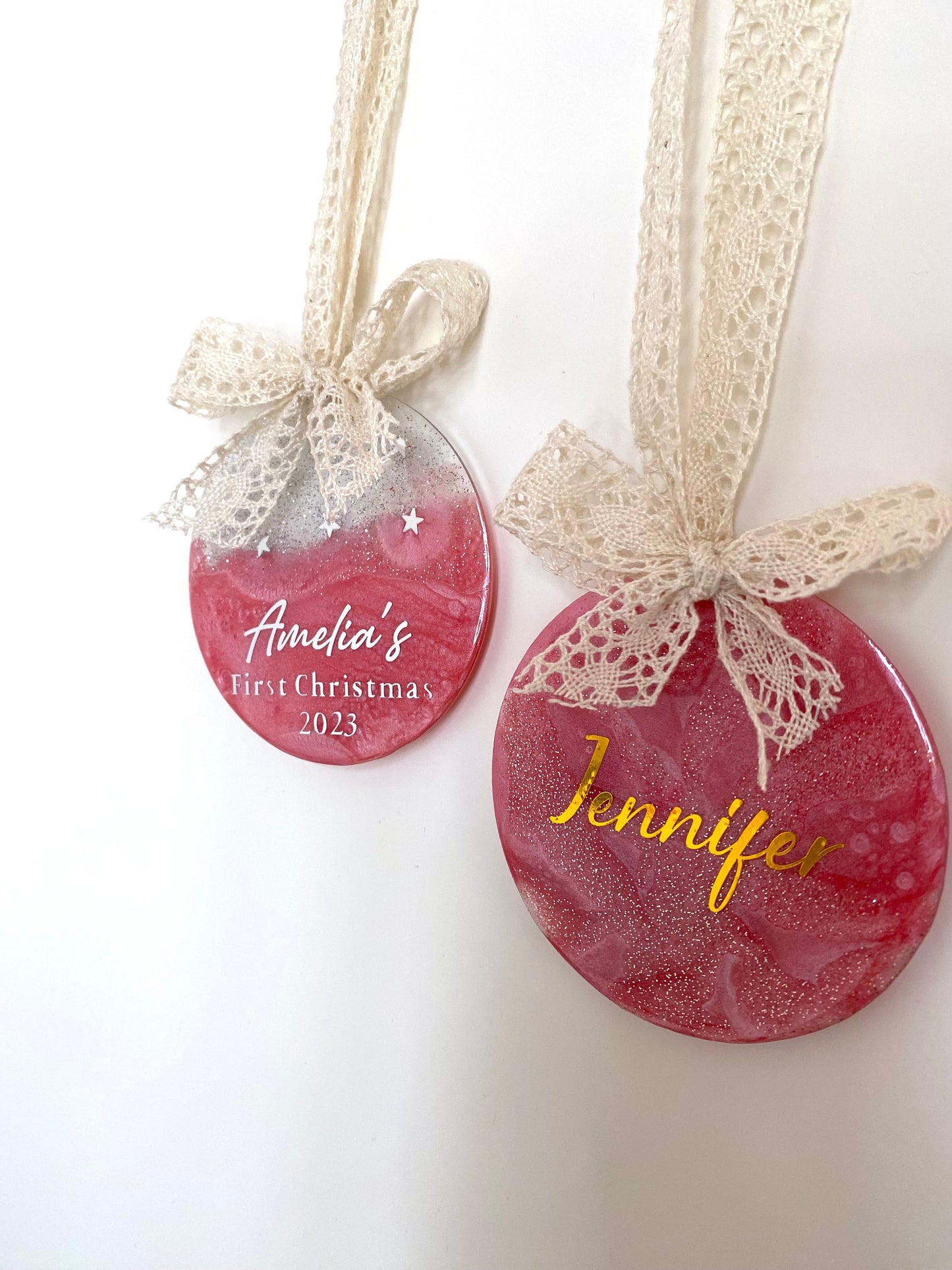 Two Pink Baby First Christmas resin baubles with glittery gold tops and twinkling stars, one personalised in silver lettering with a name, one in gold lettering with "1st Christmas", both hanging with beige lace ribbons and bows