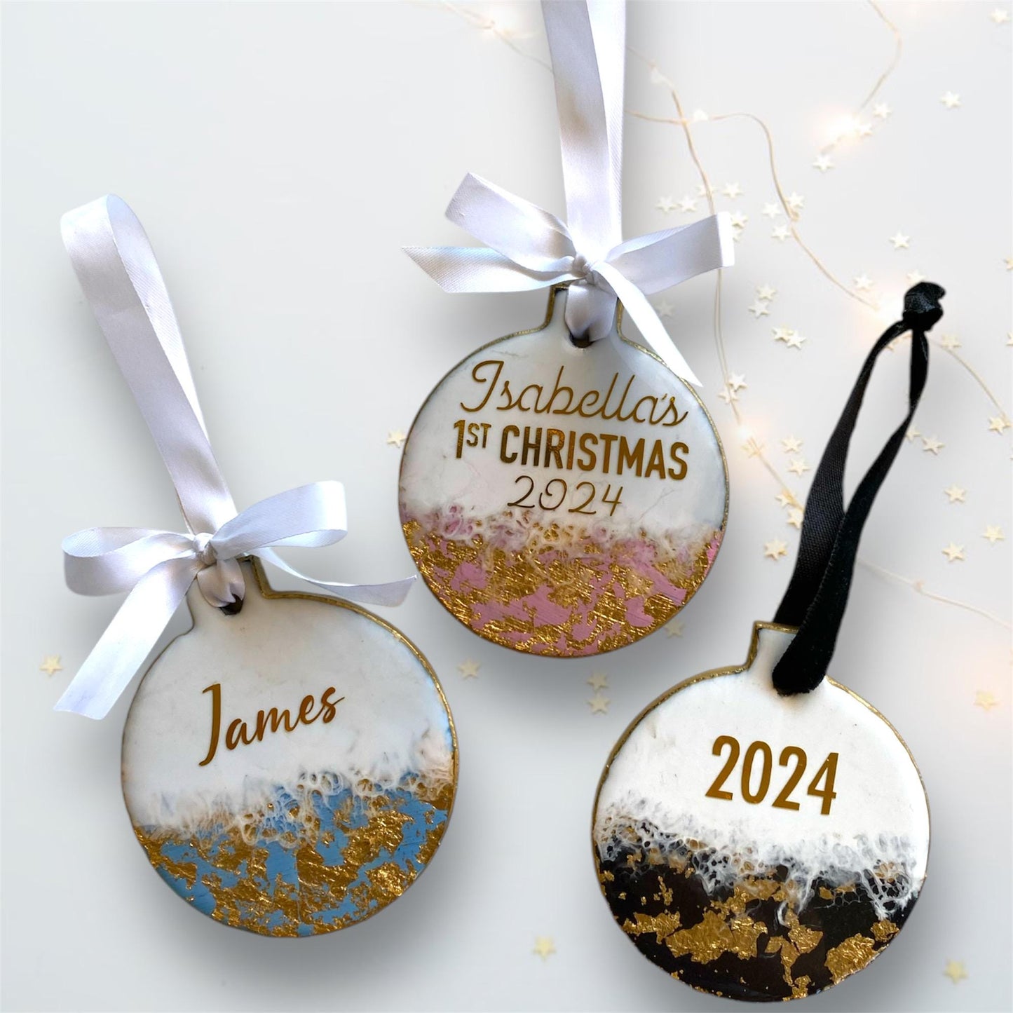 Three slate resin Baby’s First Christmas ornaments showing all personalisation options: blue with just a name, pink with baby's name and First Christmas, and black with the year 2025, all with hand-painted gold edges and white ribbons.