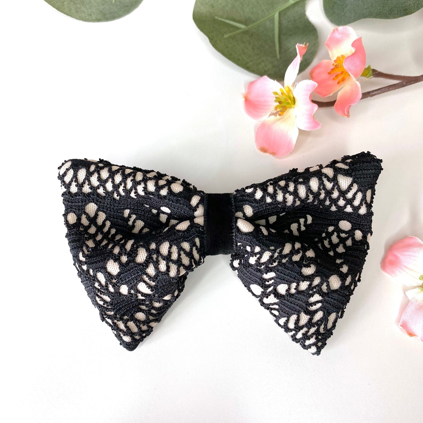 Handmade black and beige lace hair bow clips with jacquard jersey fabric and black velvet loop detail, displayed on white surface with flowers in the background