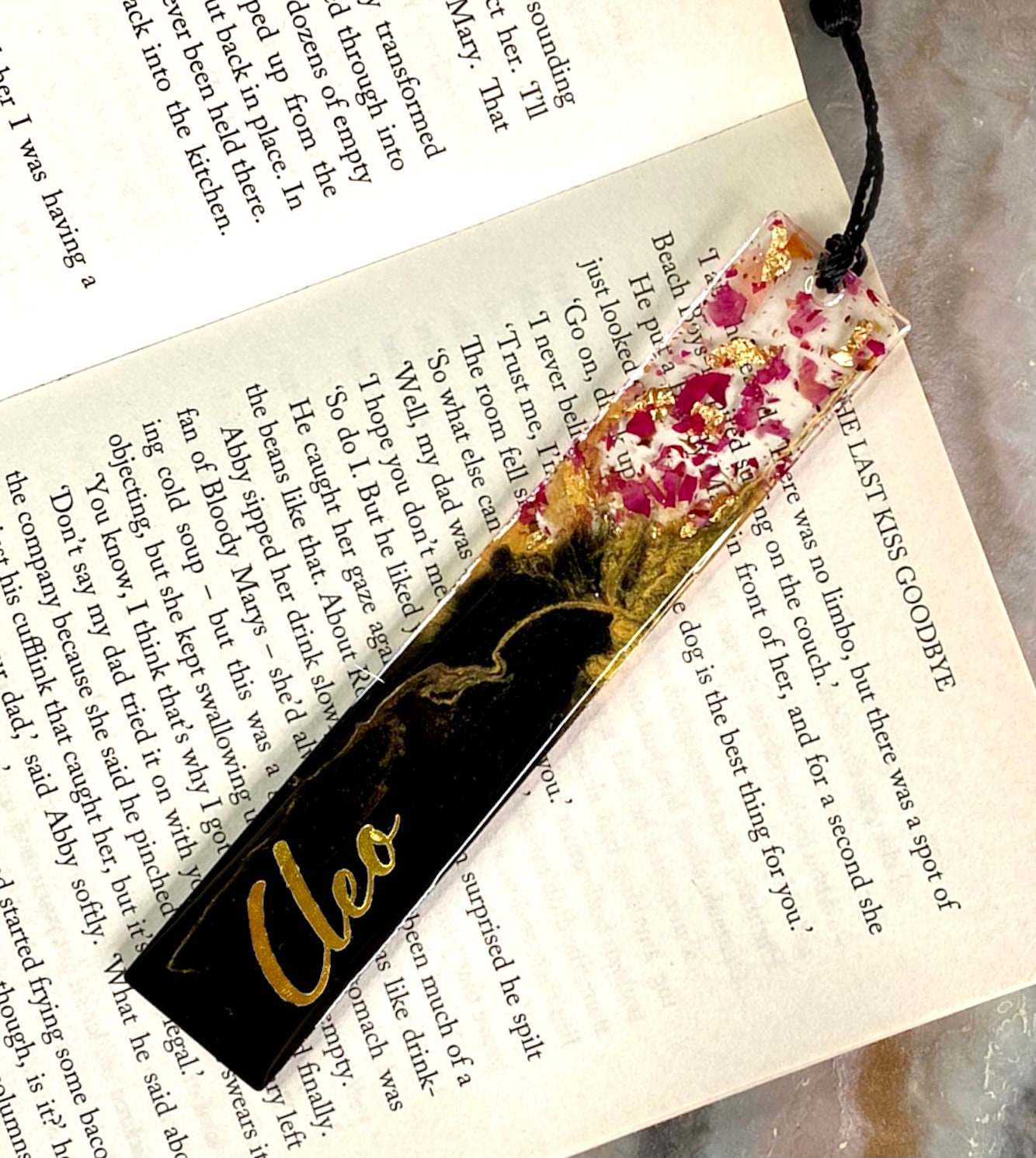 Black and gold resin bookmark personalised with gold vinyl name, placed inside a book, showing dried rose petals in clear resin