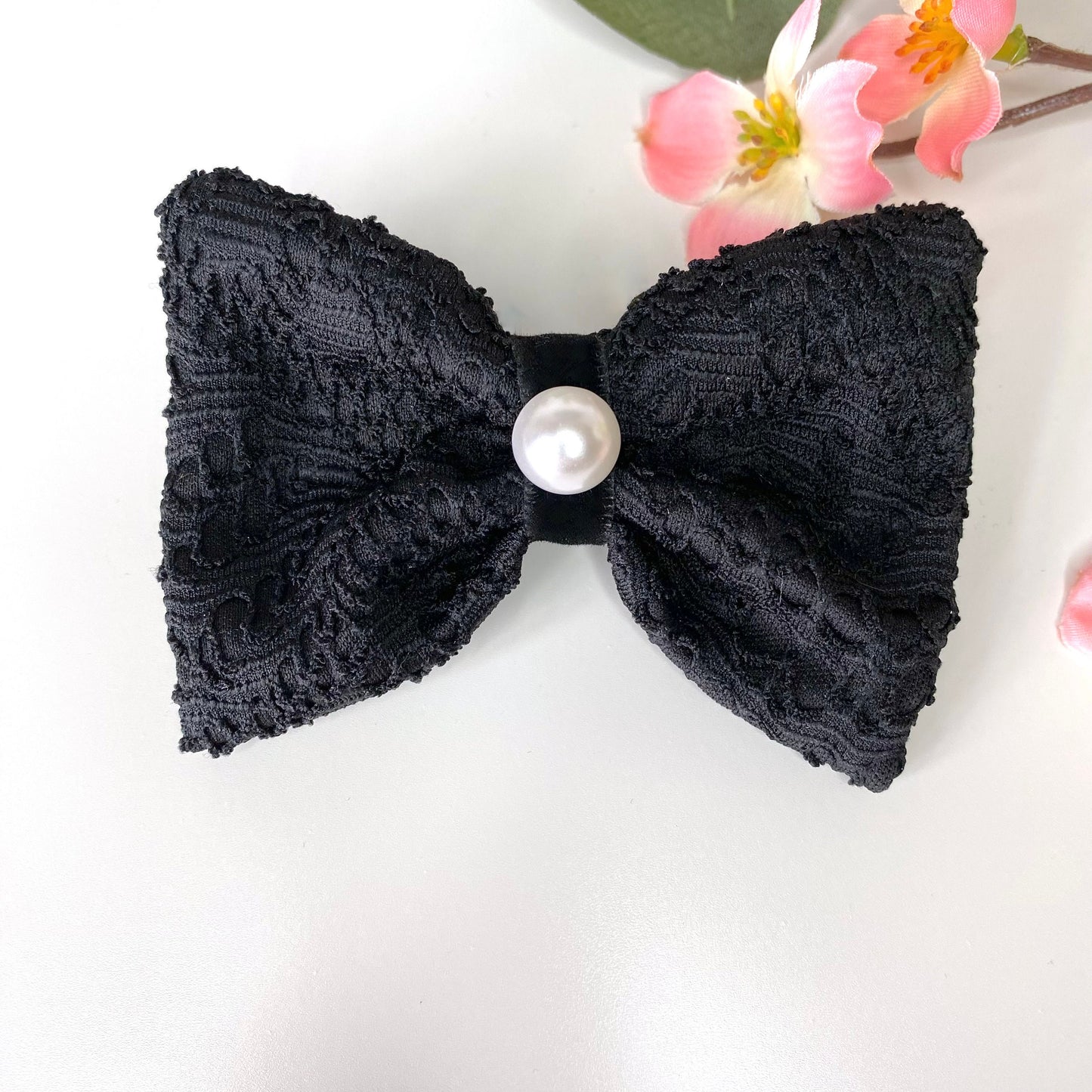 Handmade black lace hair bow clip with jacquard jersey fabric, black velvet loop, and pearl accent, displayed on white surface with flowers in the background