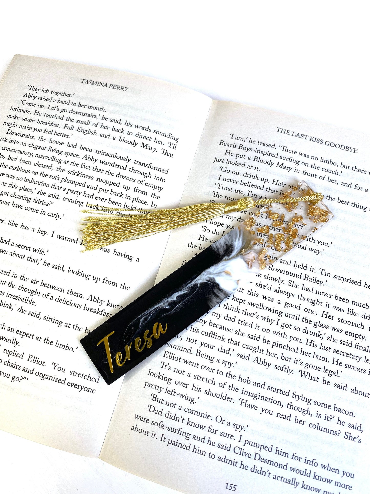 Black resin bookmark with gold leaf, personalised with gold vinyl, placed inside an open book, handmade bookmark for readers or gifts.