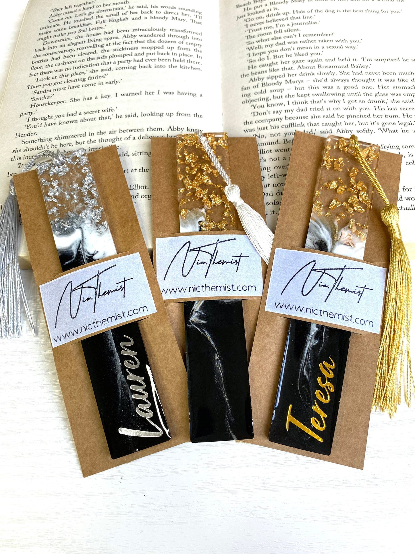 Three black and white resin bookmarks with gold and silver leaf inside packaging sleeves with logo, one personalised in gold vinyl, one personalised in silver vinyl, and one non-personalised, handmade bookmarks for readers or gifts