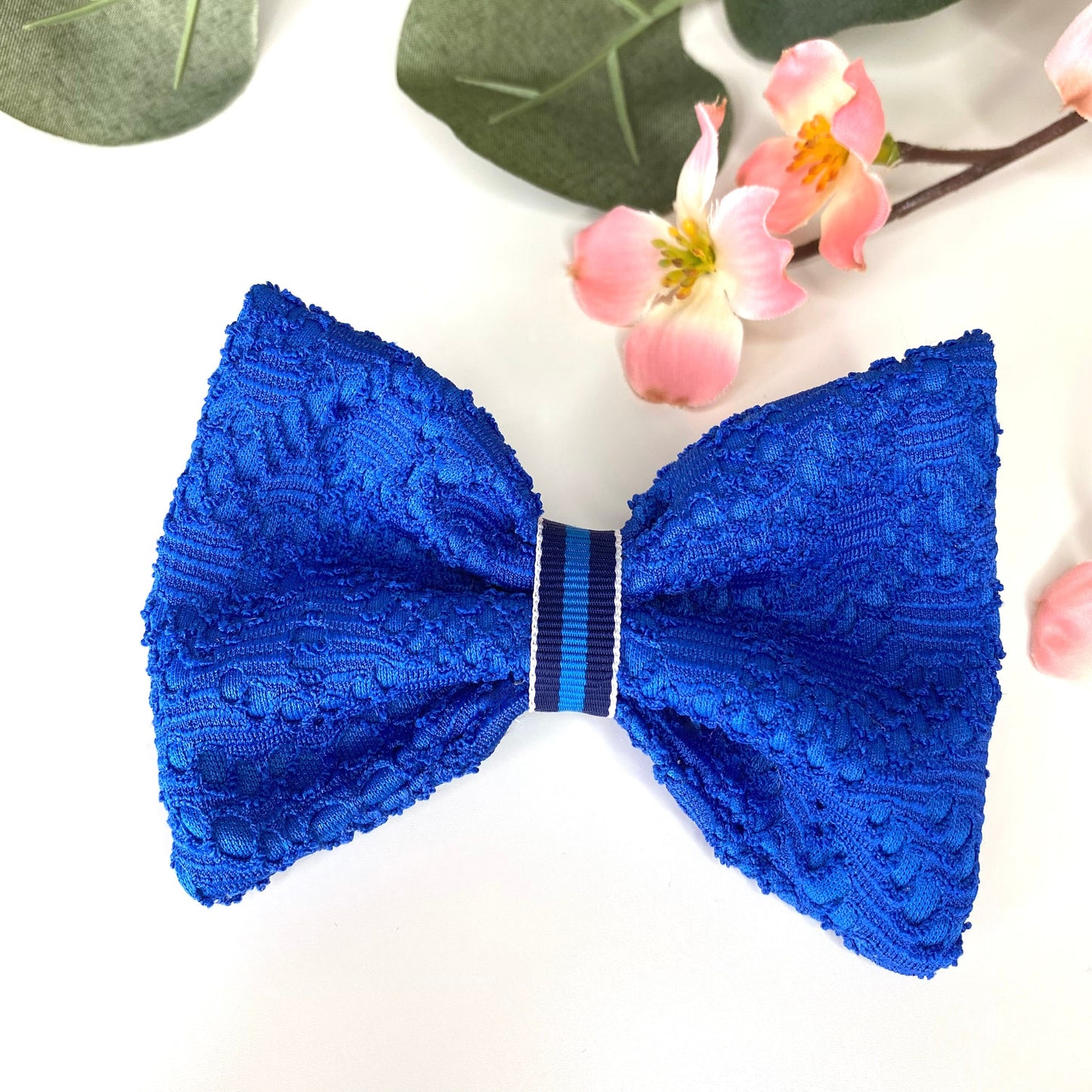 Handmade blue lace hair bow clip with jacquard jersey fabric and striped loop detail, displayed on white surface with flowers in the background