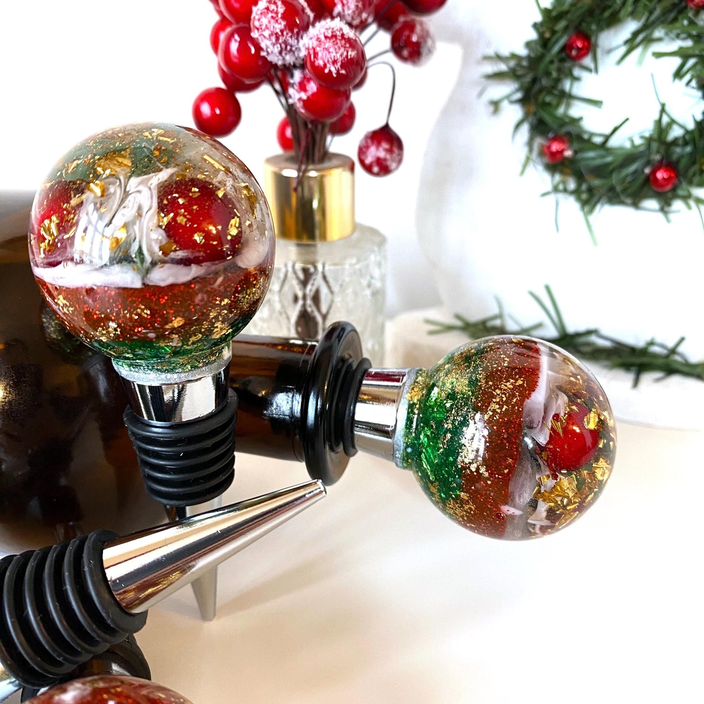Close-up of Christmas resin wine stopper in gin bottle with two additional stoppers on surface showing red berries, gold leaf, and glitter details