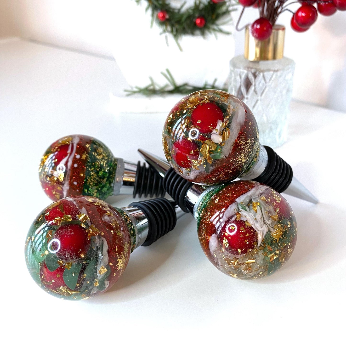 Four Christmas resin wine stoppers stacked showing red berries, gold leaf, and glitter details