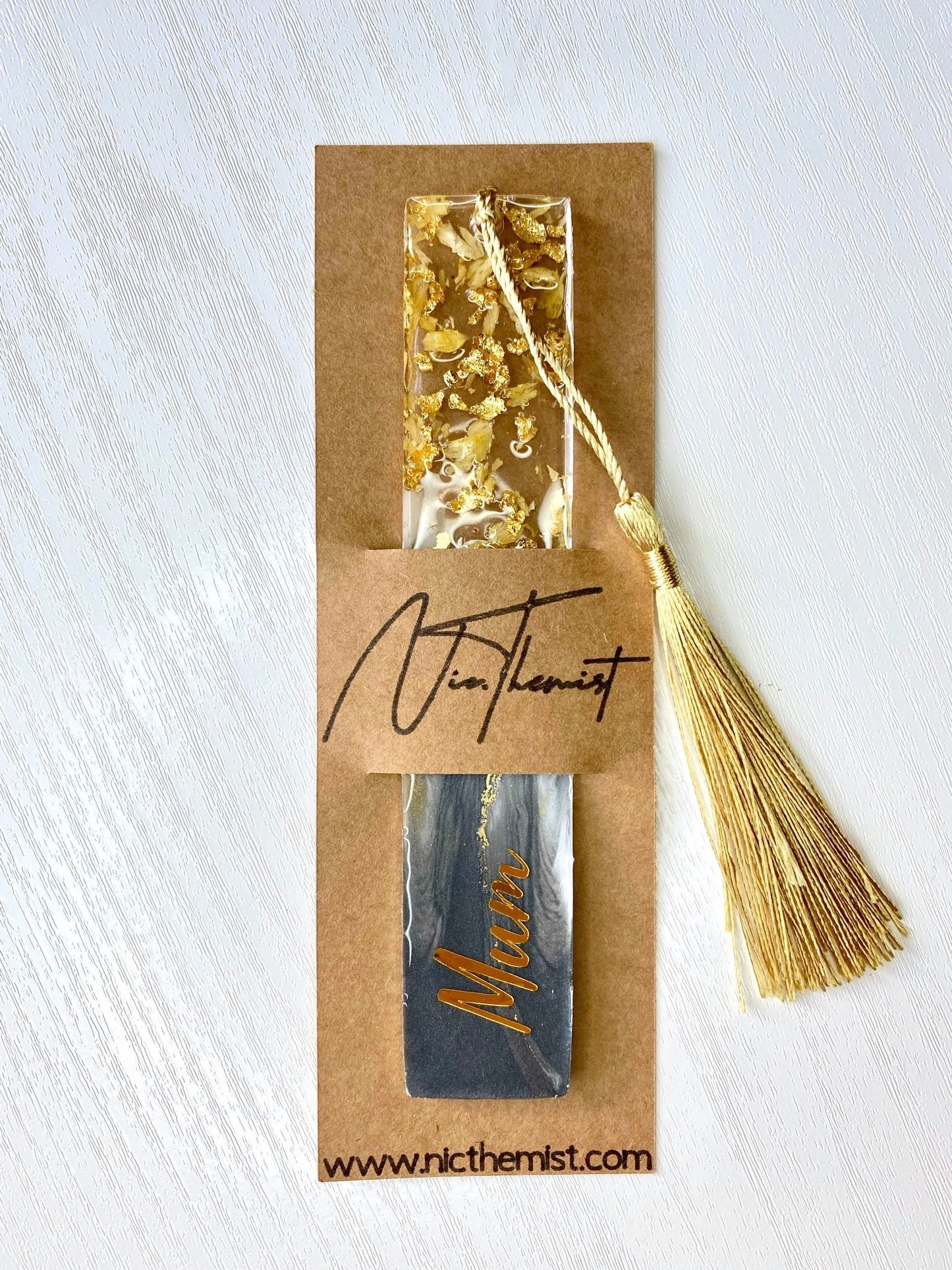 Grey resin bookmark with dried jasmine petals and gold leaf, personalised with gold vinyl, inside packaging sleeve with logo, featuring a gold tassel, handmade gift bookmark for readers