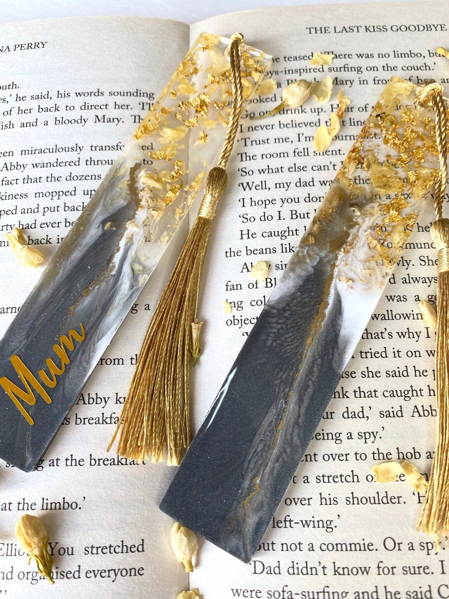 Close-up of grey resin bookmarks with dried jasmine petals and gold leaf on an open book, showing intricate handmade details and one personalised with gold vinyl.
