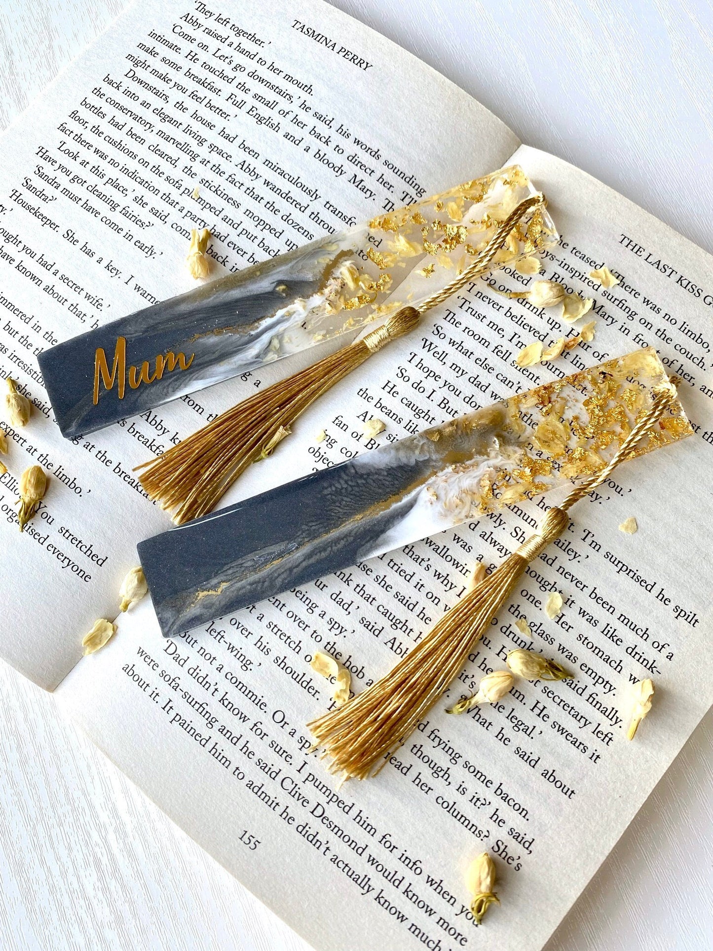 Grey resin bookmarks with dried jasmine petals and gold leaf on an open book, one personalized with gold vinyl, the other plain, elegant handmade bookmarks for gifts or reading.