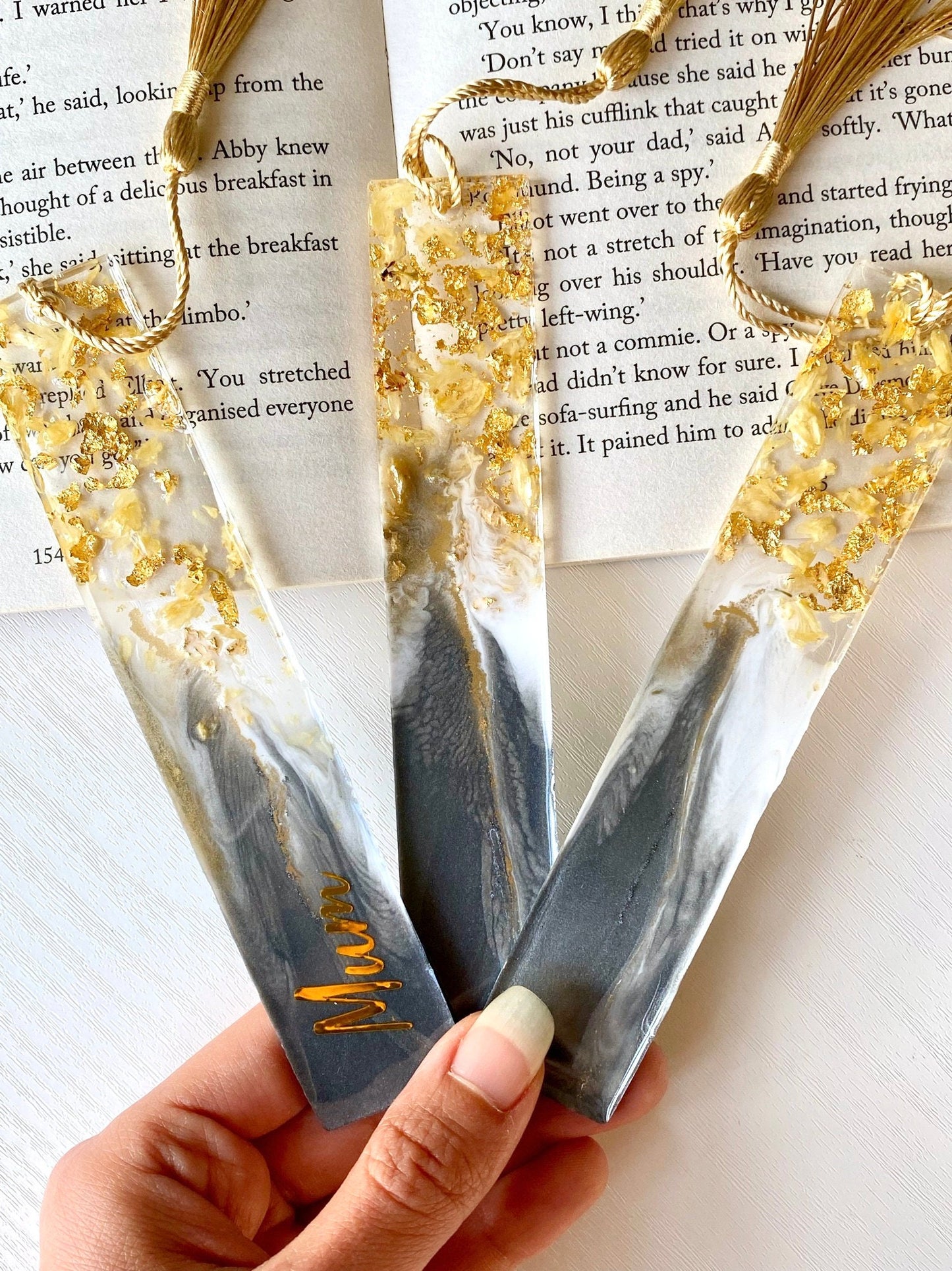 Hand holding three grey resin bookmarks with dried jasmine petals and gold leaf, one personalised with gold vinyl and two plain, elegant handmade bookmarks for gifts or reading.