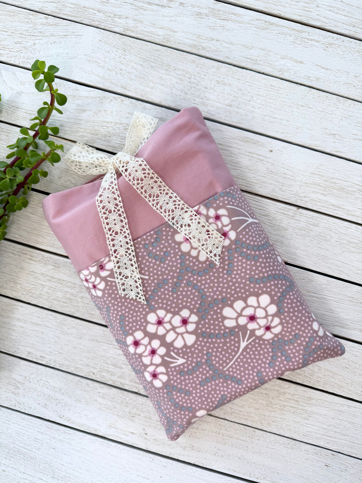 Handmade dusty pink fabric book pouch laid flat on a white wooden surface, showing the contrasting floral front pocket and cream lace ribbon closure.