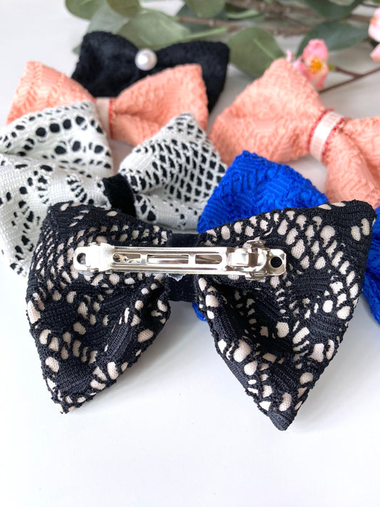 Close-up of handmade lace hair bow clip upside down showing silver spring barrette fastening, with blurred clips in the background on white surface