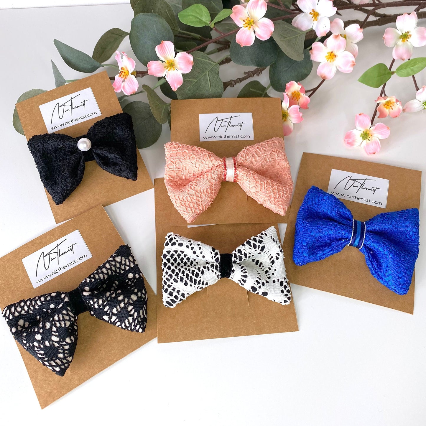 Handmade lace hair bow clips in black, blue, white, and peach attached to branded packaging sleeves, displayed on white surface with flowers in background