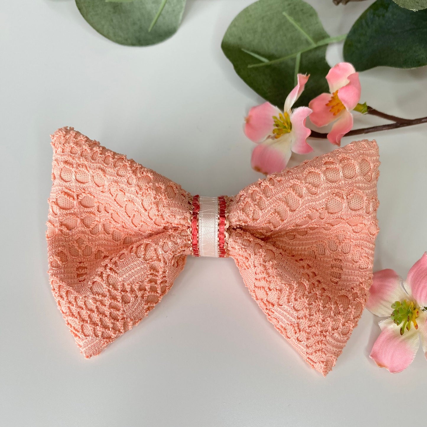 Handmade peach lace hair bow clip with jacquard jersey fabric and satin strip loop detail, displayed on white surface with flowers in the background