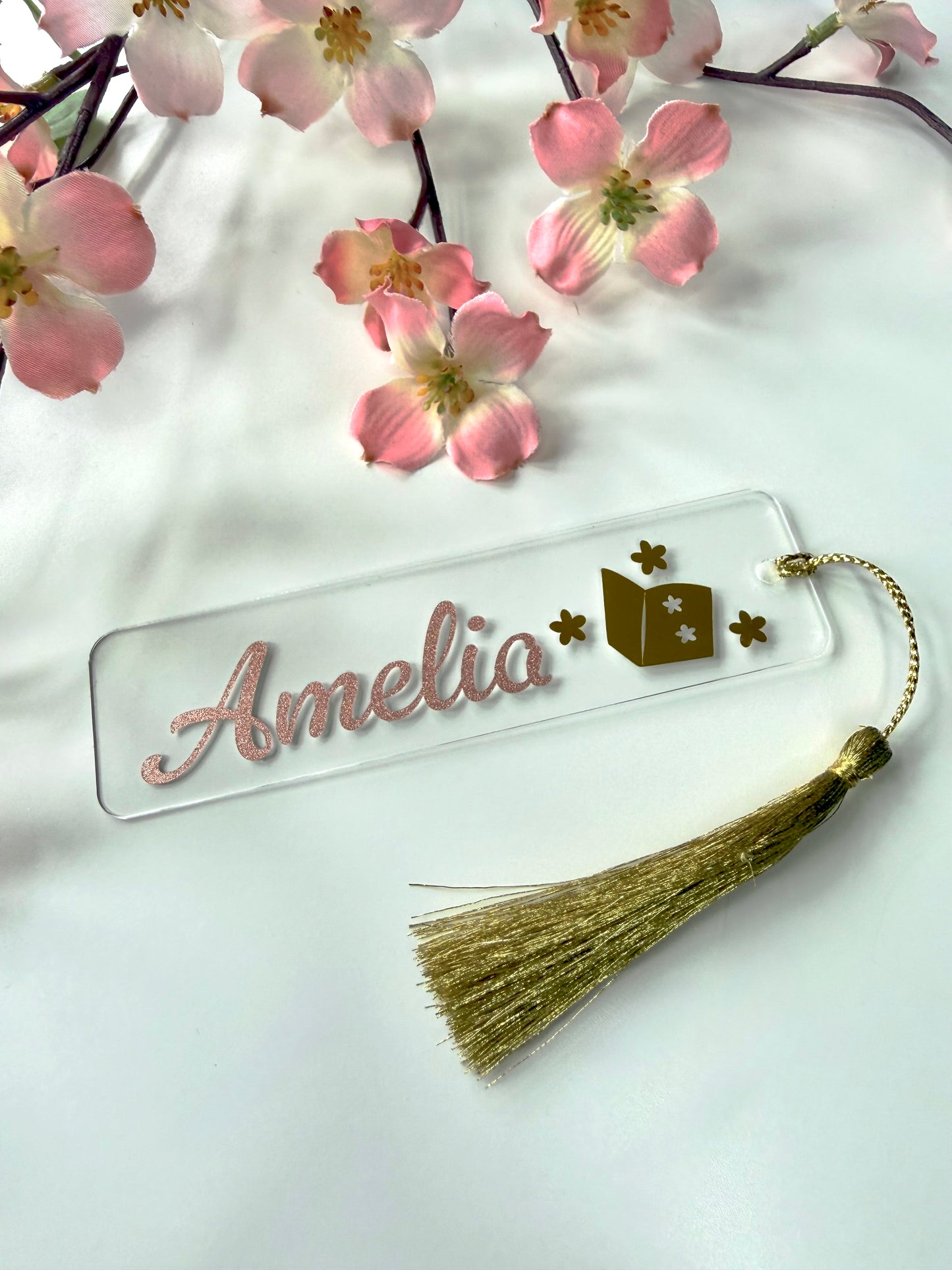 Personalised acrylic bookmark with rose gold glitter name and open book design, lying flat on a white surface with a matching tassel.