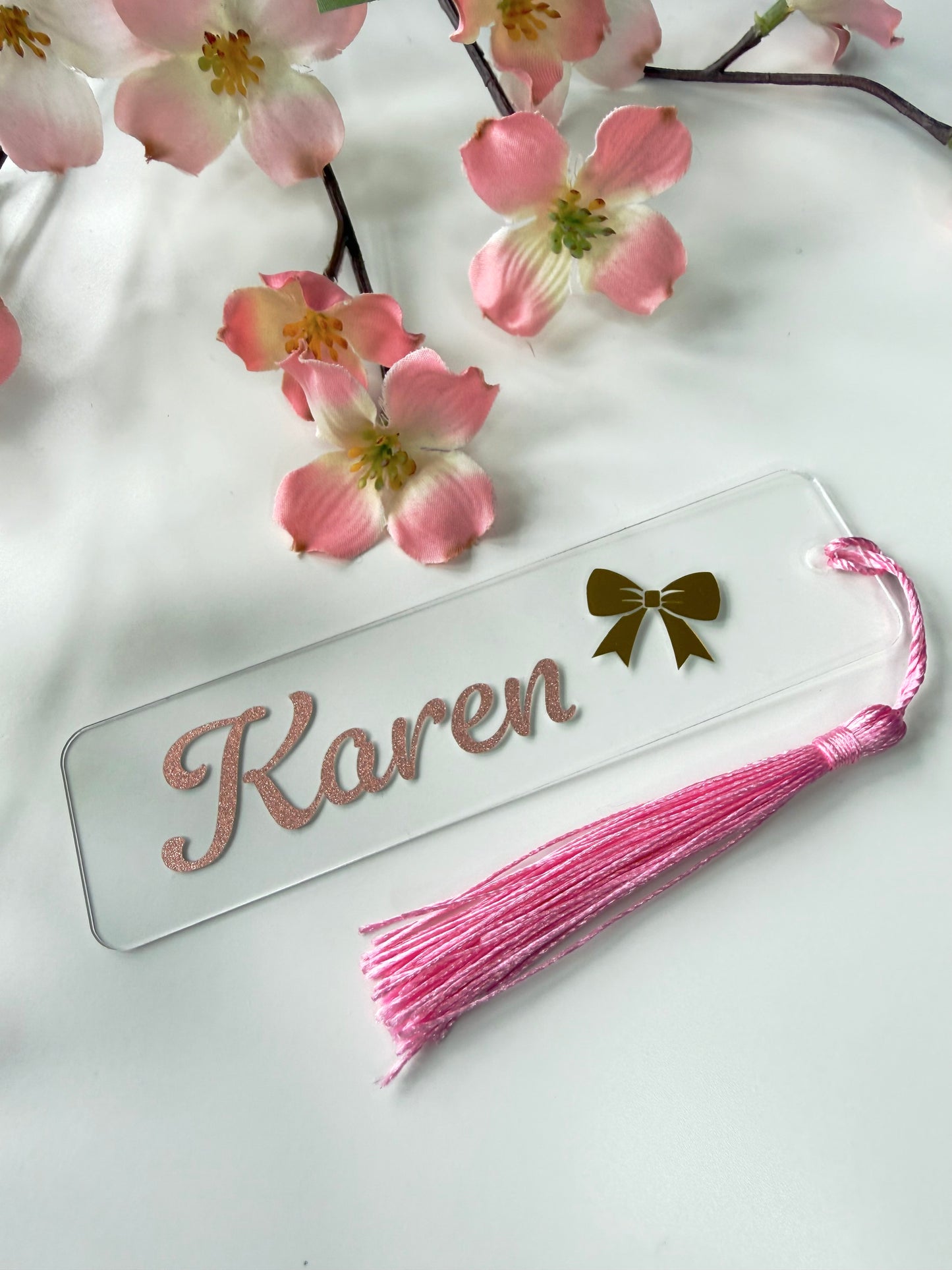 Personalised acrylic bookmark with rose gold glitter name and bow design, lying flat on a white surface with a matching tassel.