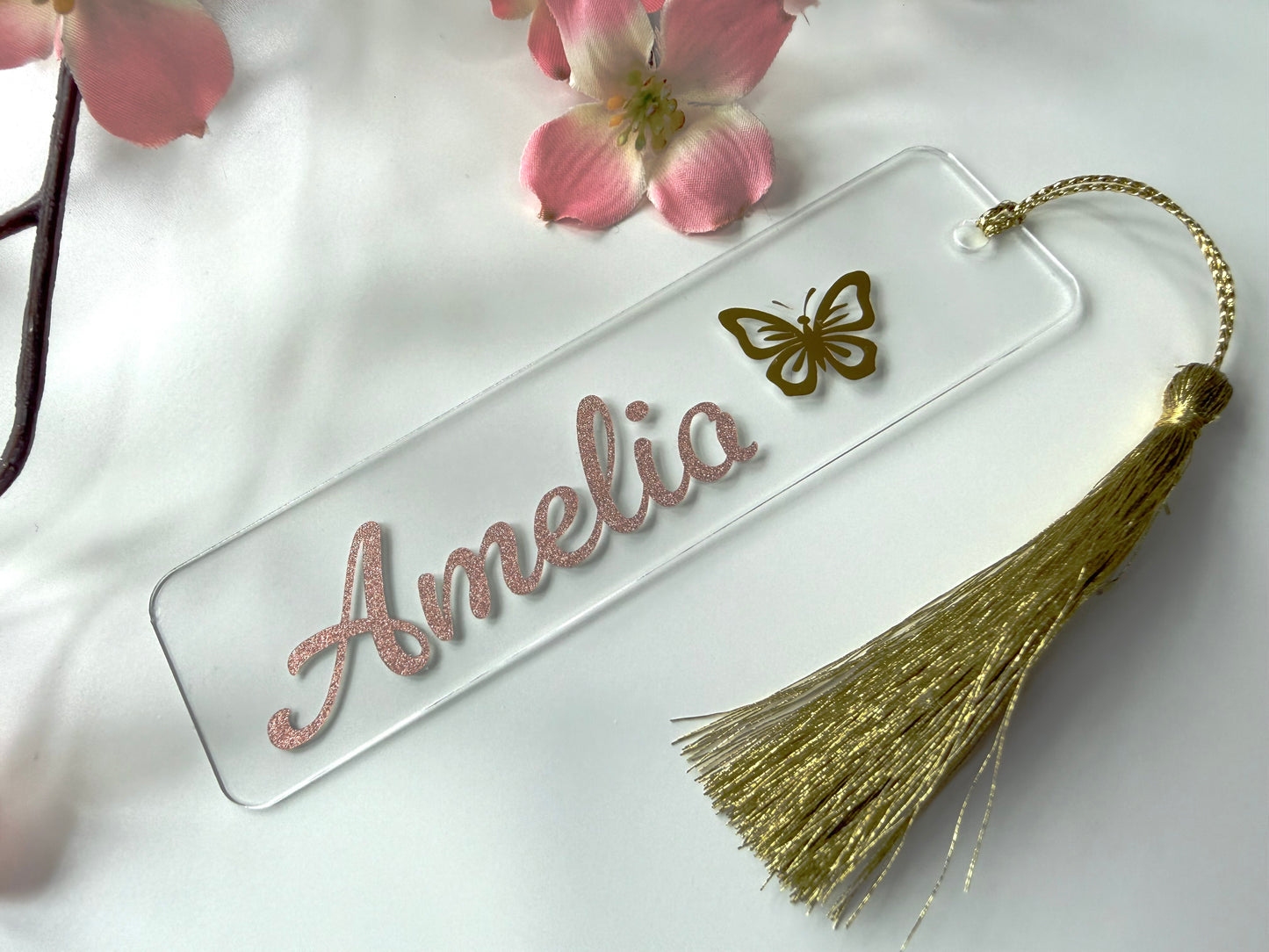 Personalised acrylic bookmark with rose gold glitter name and butterfly design, lying flat on a white surface with a matching tassel.