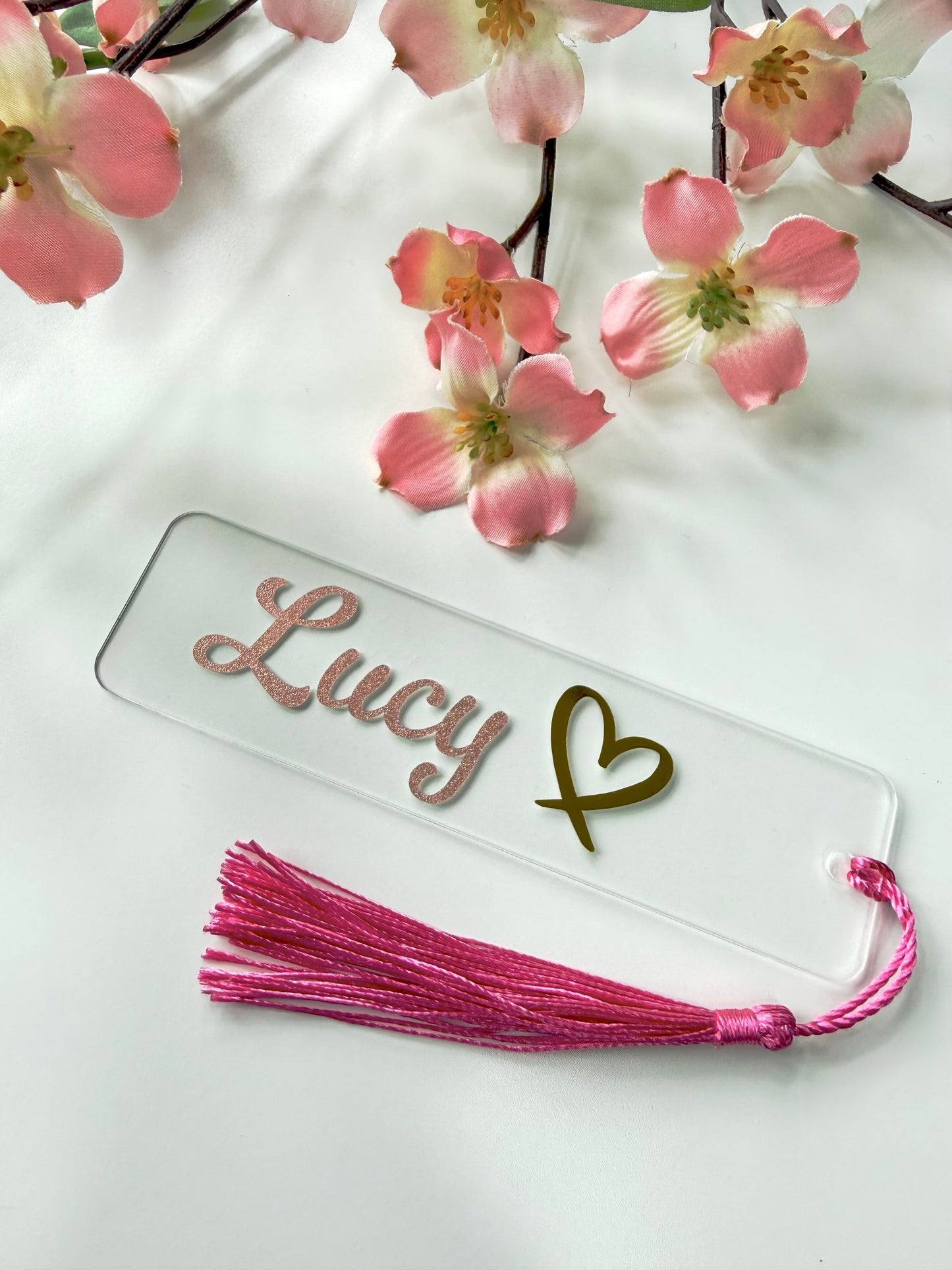 Personalised acrylic bookmark with rose gold glitter name and heart design, lying flat on a white surface with a matching tassel.