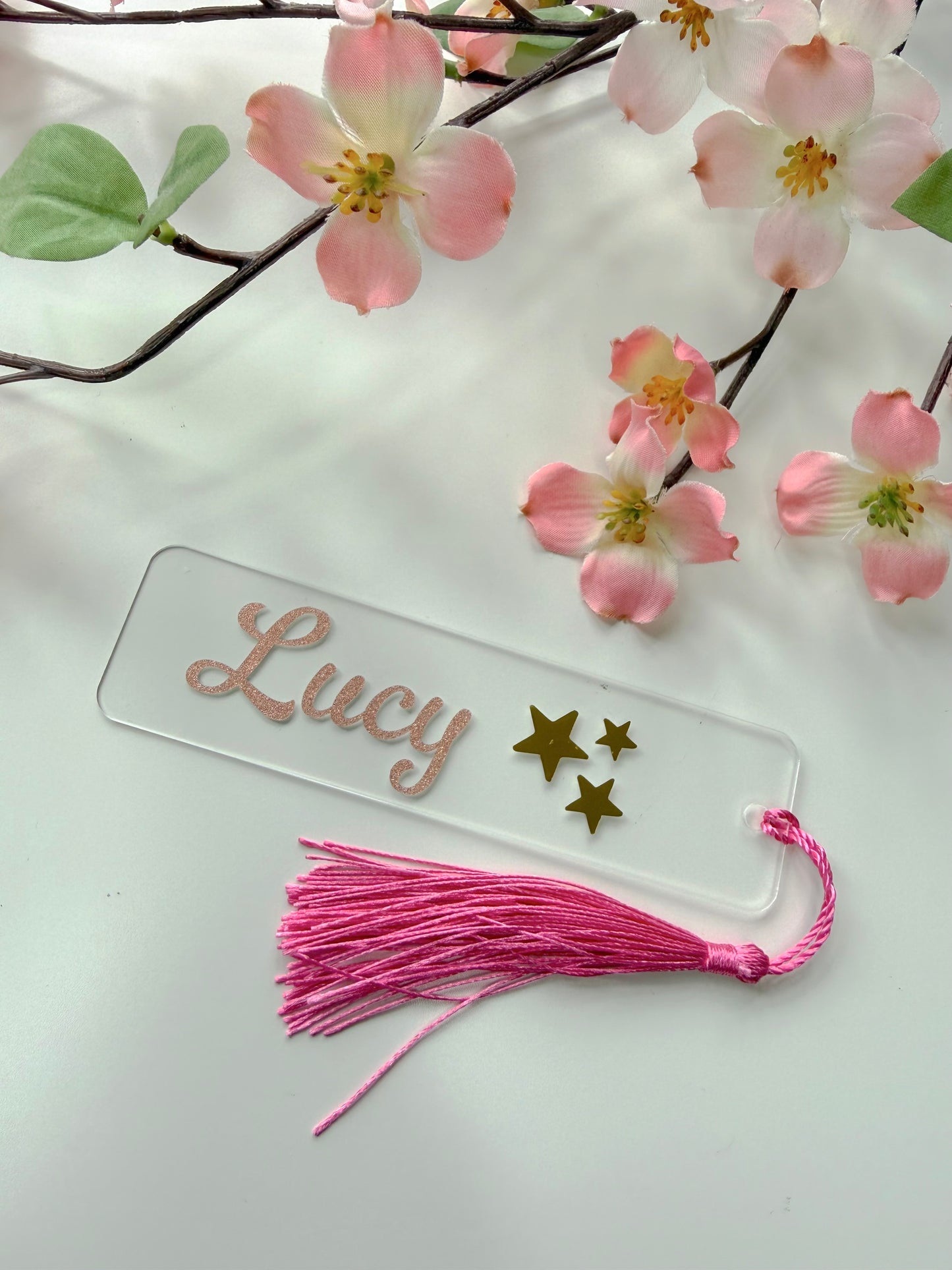 Personalised acrylic bookmark with rose gold glitter name and stars design, lying flat on a white surface with a matching tassel.