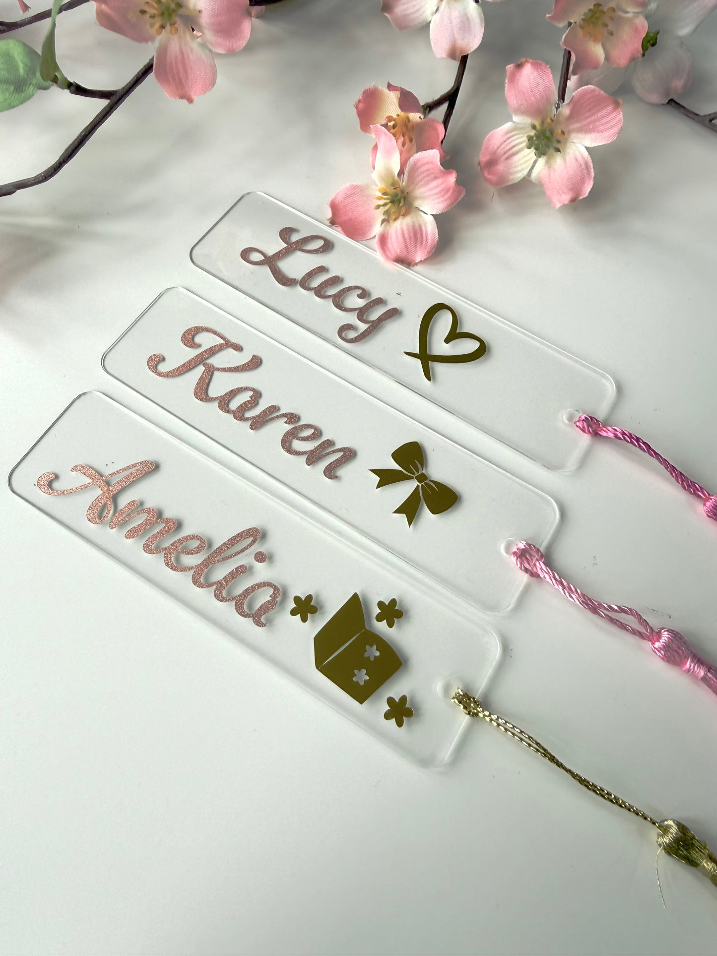 Three personalised acrylic bookmarks in rose gold glitter with bow, heart, and book designs, lying flat on a white surface with matching tassels.
