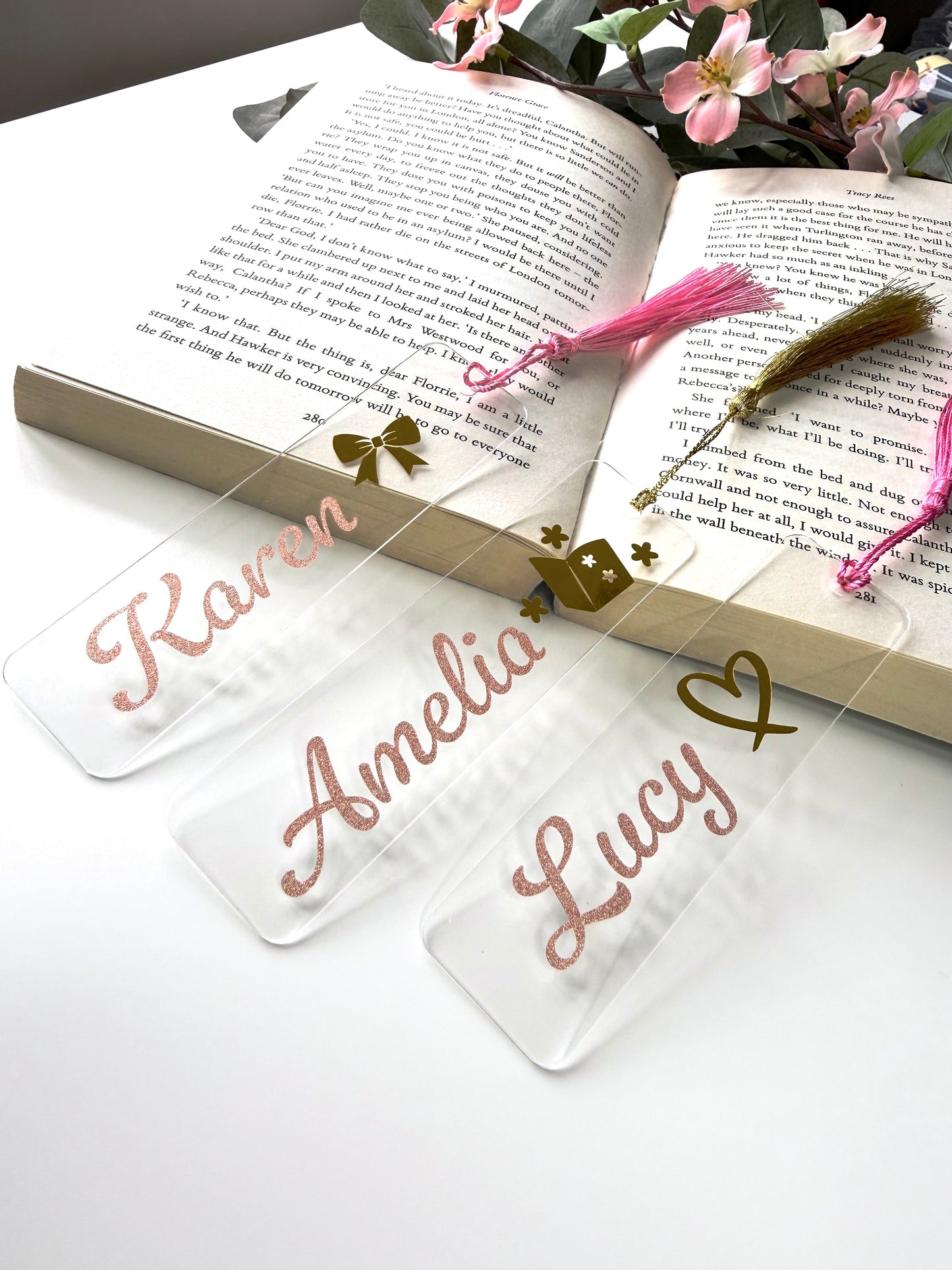 Three personalised acrylic bookmarks in rose gold glitter with bow, heart, and book designs, displayed on an open book with the bottoms resting on a white surface and flowers in the background.