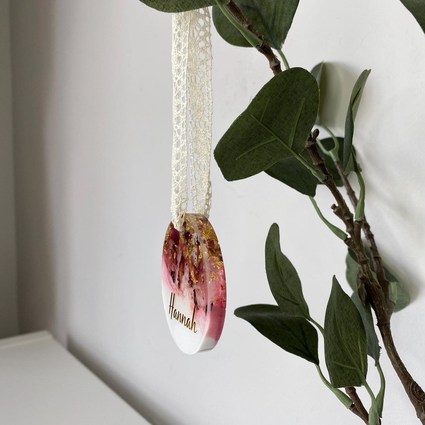 Personalised resin Christmas ornament with dried rose petals and gold leaf, angled to show thickness, custom name in gold vinyl, pink and white colours, lace ribbon included.