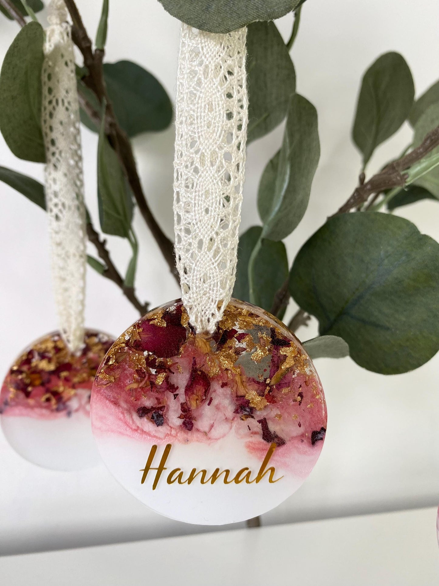Two resin Christmas ornaments with dried rose petals and gold leaf hanging on foliage; front ornament personalised with a custom name in gold vinyl, back ornament slightly blurred, pink and white colours, lace ribbons included.