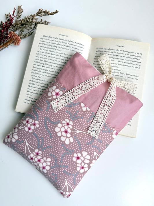 A pink fabric pouch with a white floral pattern and a cream lace ribbon bow, styled on top of an open book with dried flowers in the corner.