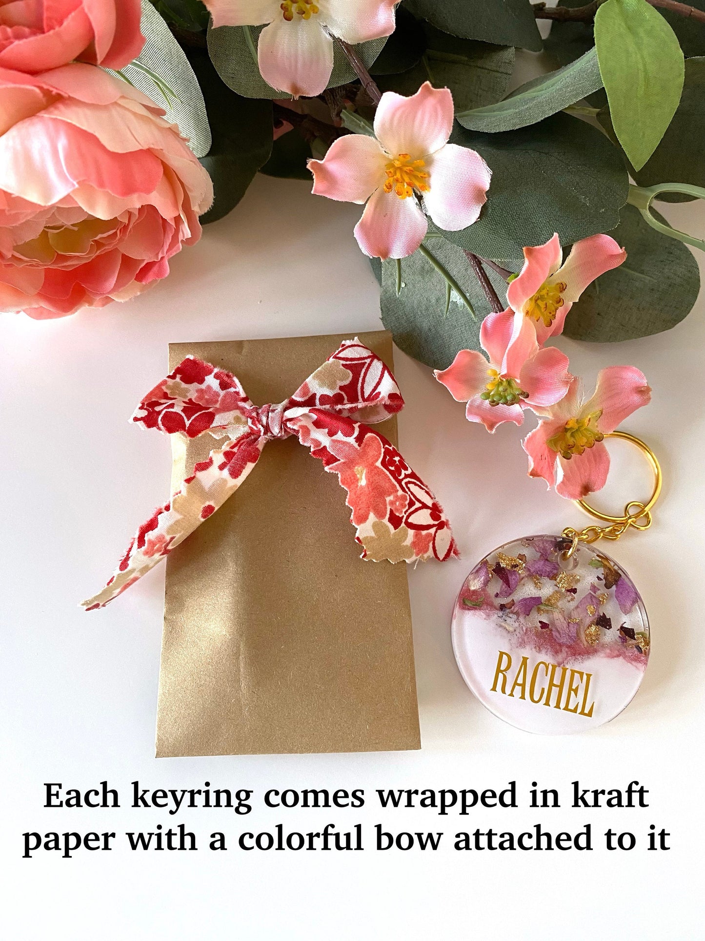 Personalised pink and white resin keychain with dried flowers and gold leaf next to Kraft paper packaging with colourful fabric bow, ready to gift, handmade keyring for presents