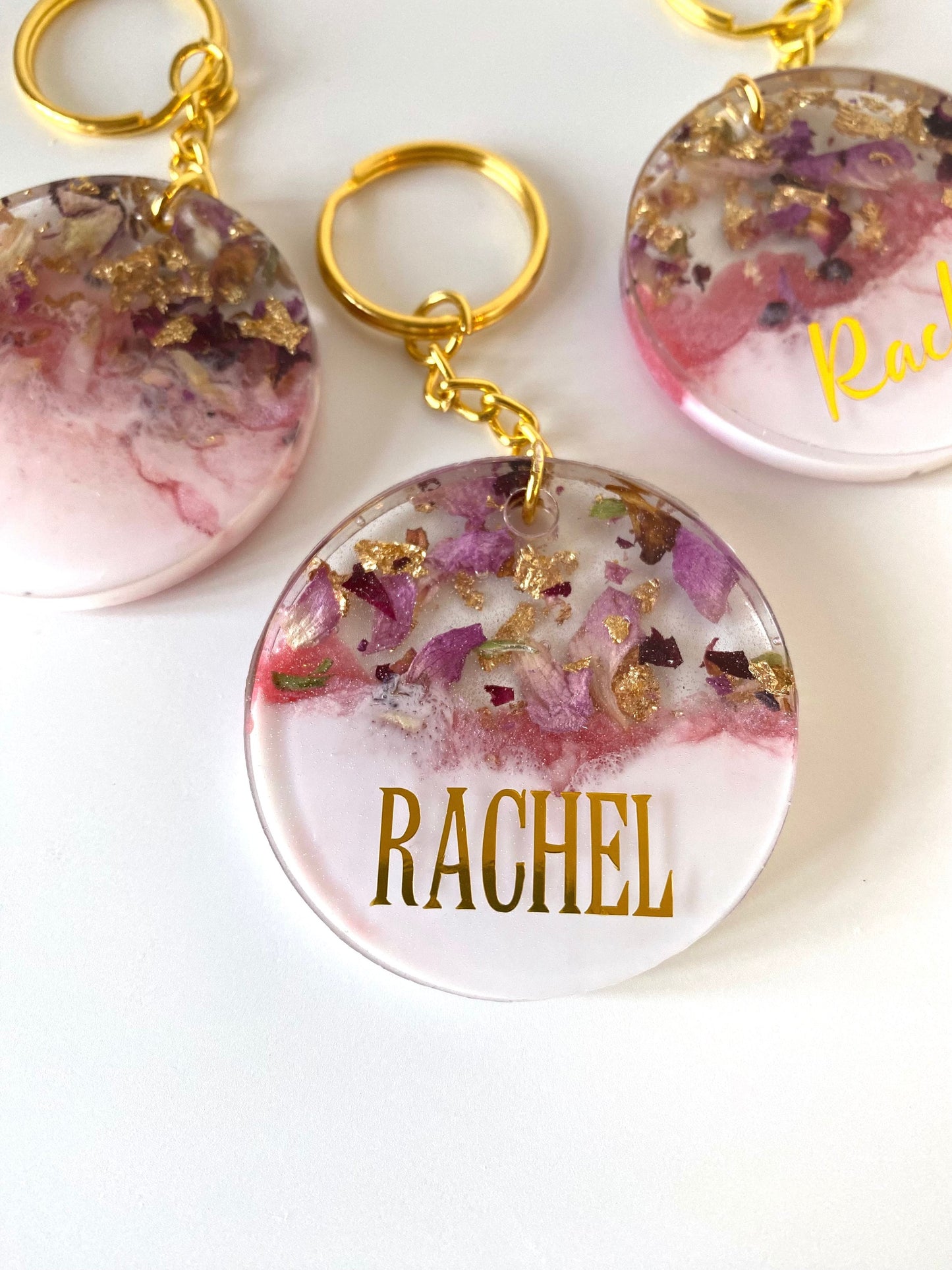 Close-up of pink and white round resin keychain with dried flowers and gold leaf, personalised in uppercase gold vinyl font, elegant handmade keyring for gifts
