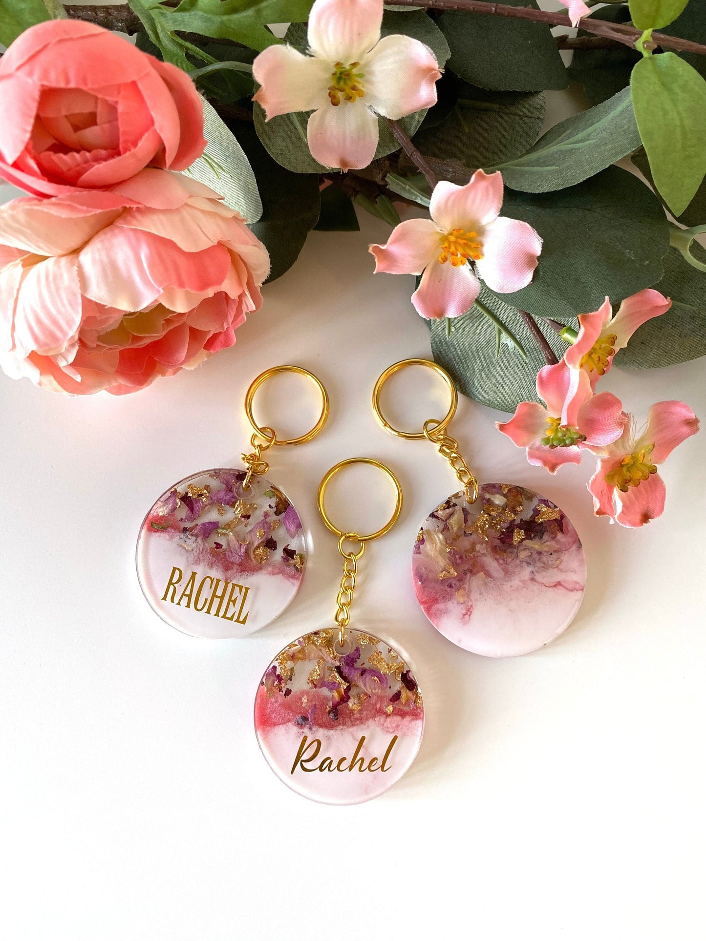 Pink and white round resin keychains with dried flowers and gold leaf, two personalised with gold vinyl names and one plain, handmade personalised keyrings for gifts