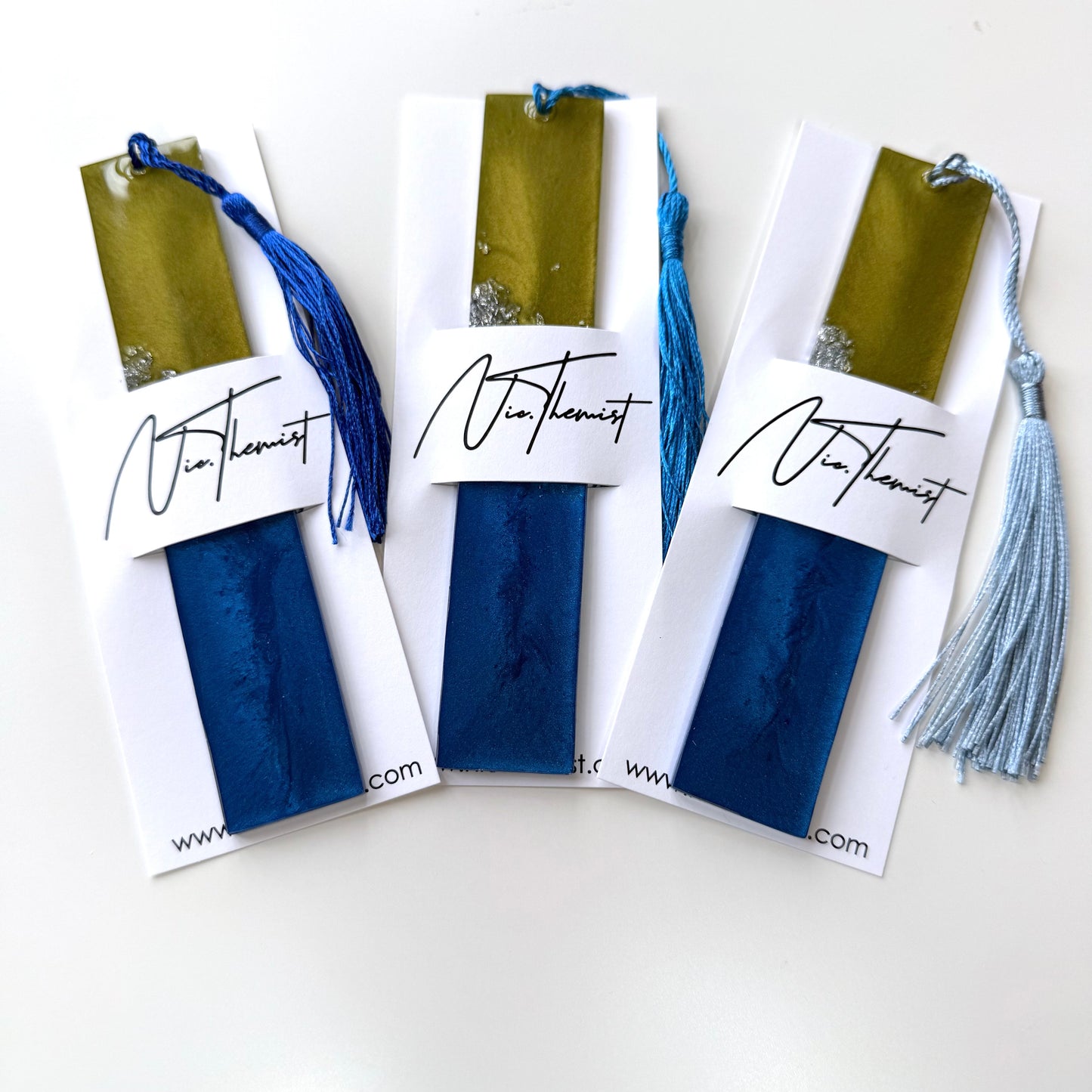 Three plain handmade resin bookmarks in branded packaging sleeve with NicThemistTreasures logo. Blue, green, and silver celestial-inspired design, perfect gift for book lovers