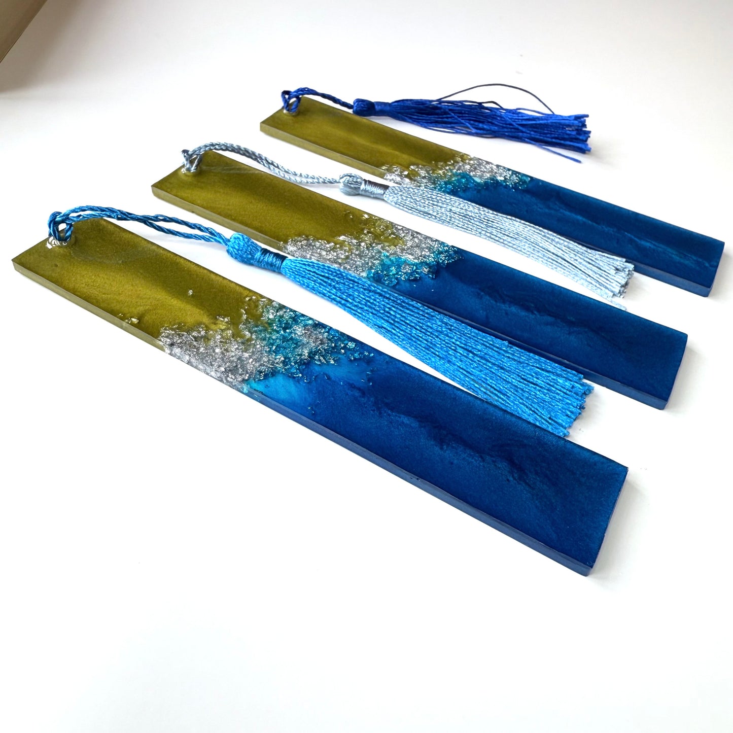 “Three plain handmade resin bookmarks with tassels on white surface, angled to show thickness. Blue, green, and silver celestial-inspired design, ideal gift for book lovers.