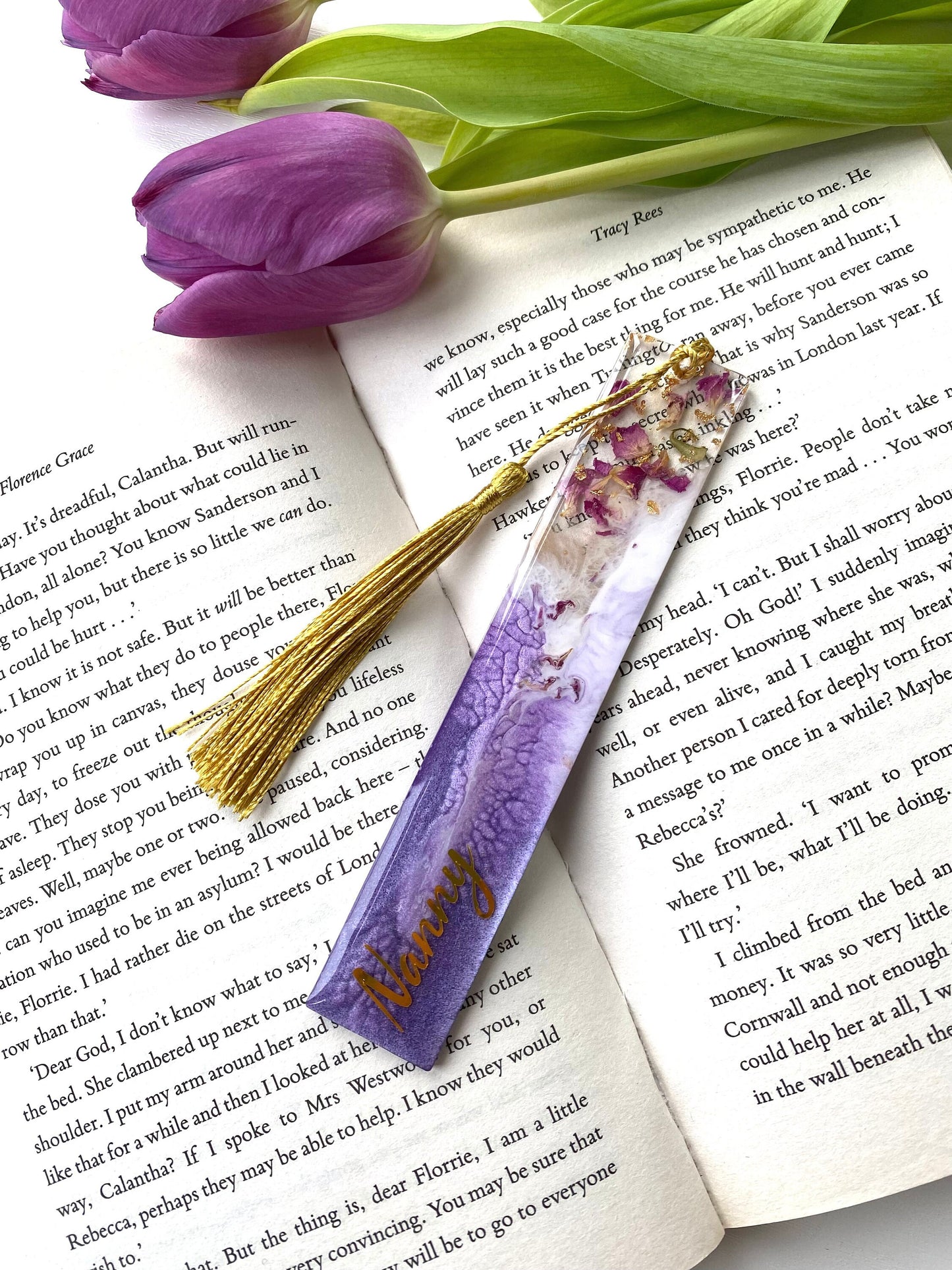 Purple resin bookmark with dried roses and gold leaf, personalised with gold vinyl, placed inside an open book, handmade bookmark for readers or gifts.