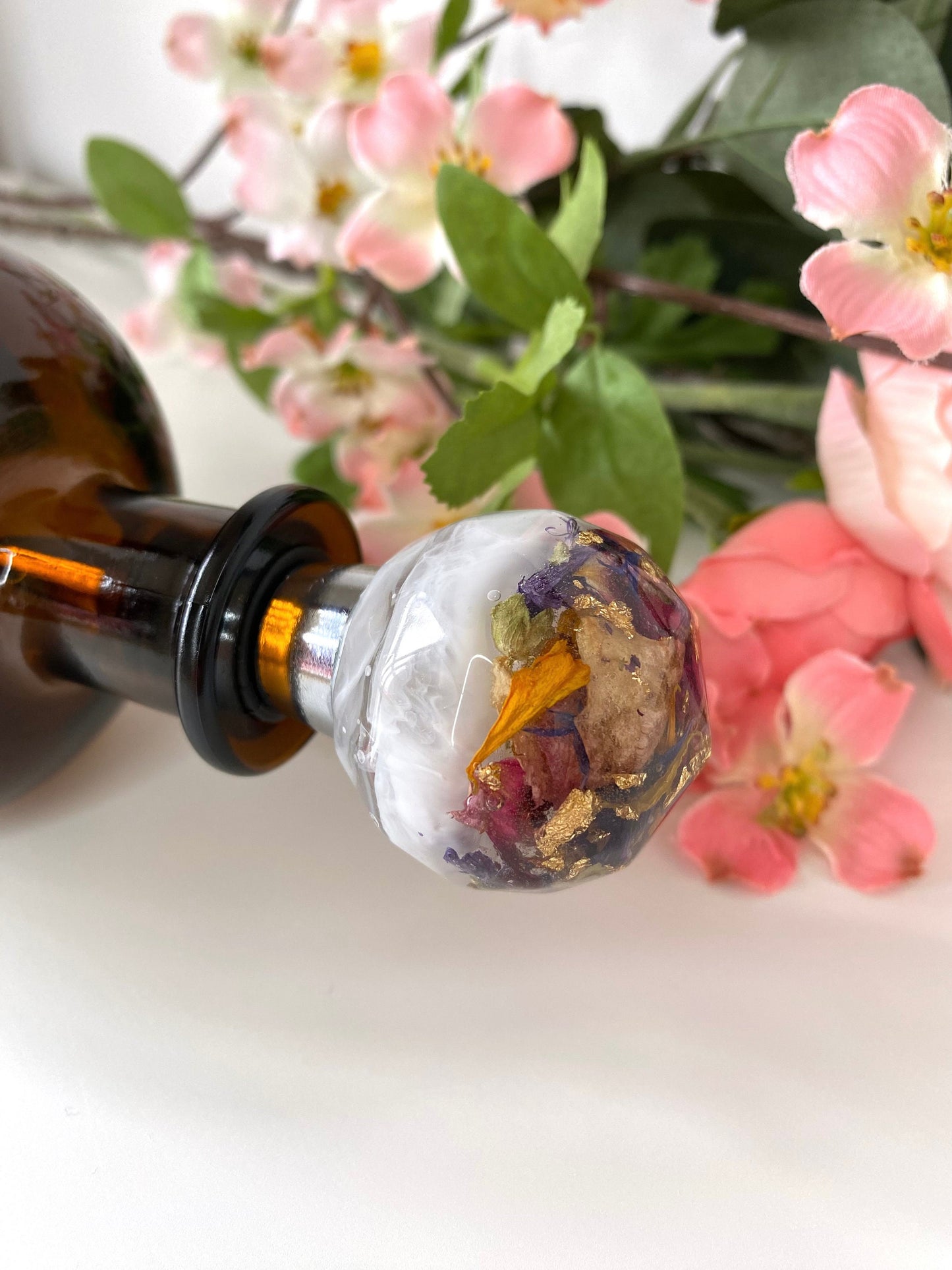 Close-up of handmade resin bottle stopper with stainless steel base, white resin top with clear resin containing colourful dried flowers and gold leaf, inserted into the mouth of a bottle lying flat on table, decorative and functional wine accessory for gifts or home use.