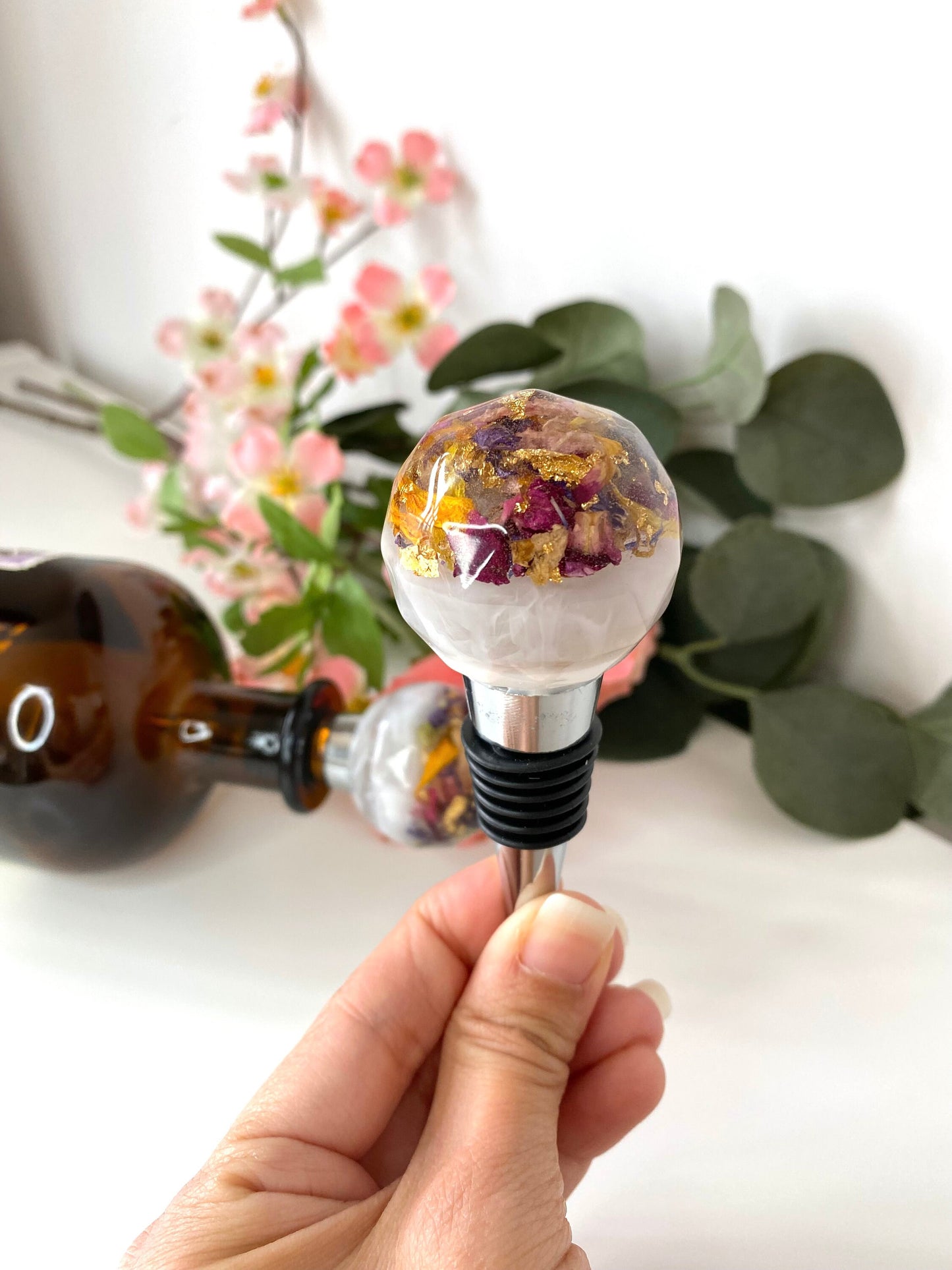 Hand holding a handmade resin bottle stopper with stainless steel base, white resin top with clear resin containing colourful dried flowers and gold leaf, decorative and functional wine accessory for gifts or home use