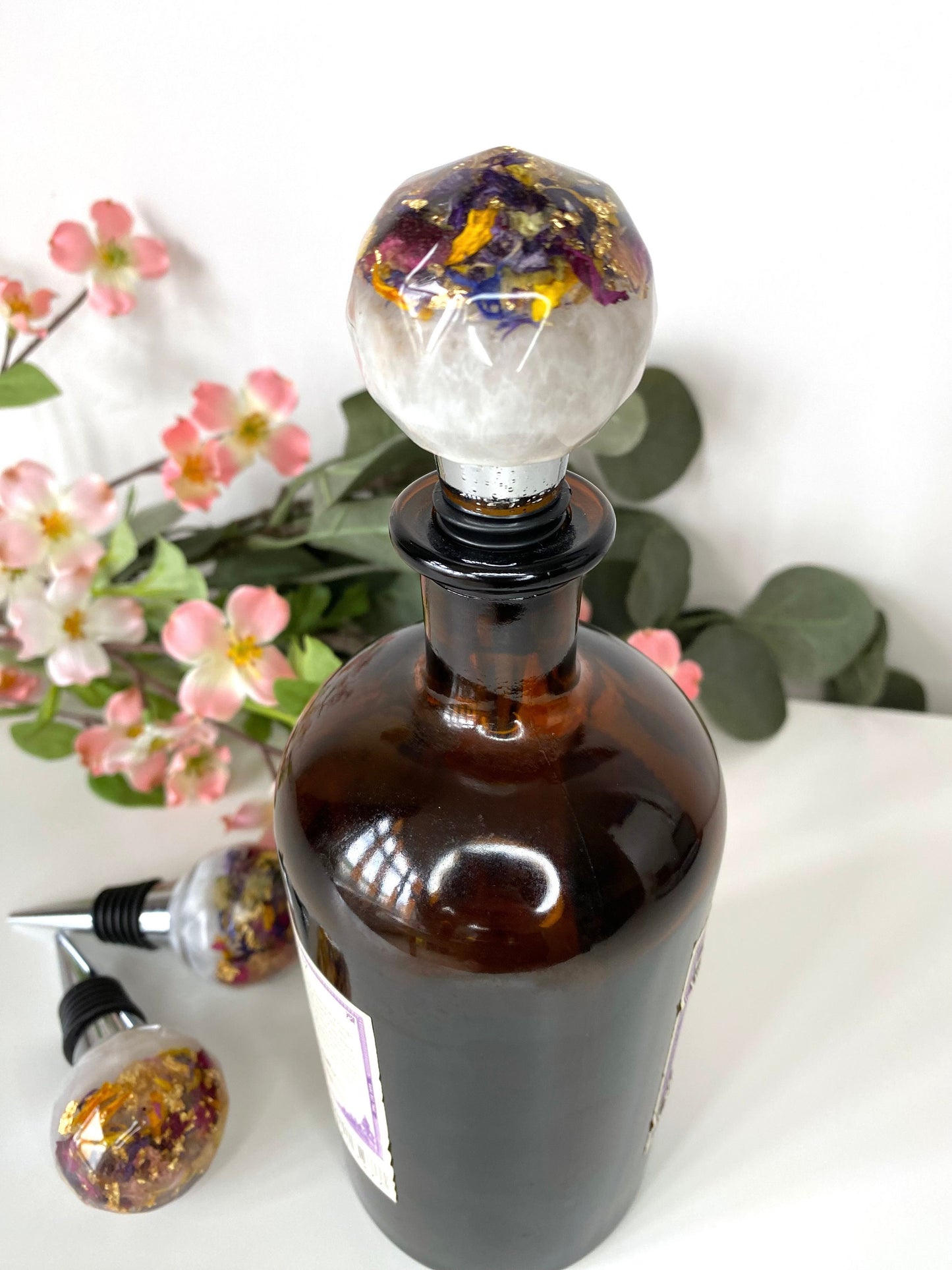 Handmade resin bottle stopper with stainless steel base, white resin top with clear resin containing colourful dried flowers and gold leaf, inserted into a bottle on a table, decorative and functional wine accessory for gifts or home use.