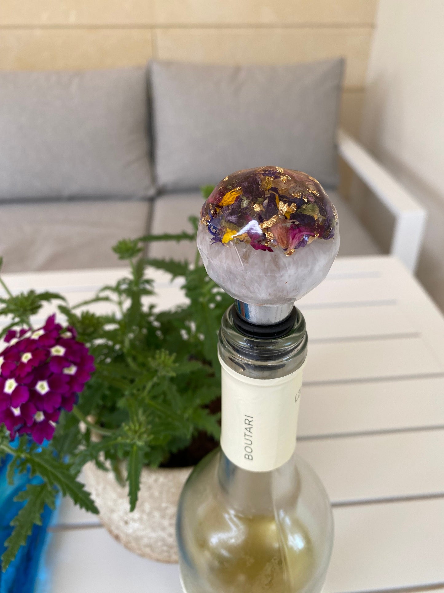Handmade resin bottle stopper with stainless steel base, white resin top with clear resin containing colourful dried flowers and gold leaf, inserted into a wine bottle on outdoor table, decorative and functional wine accessory for gifts or home use.