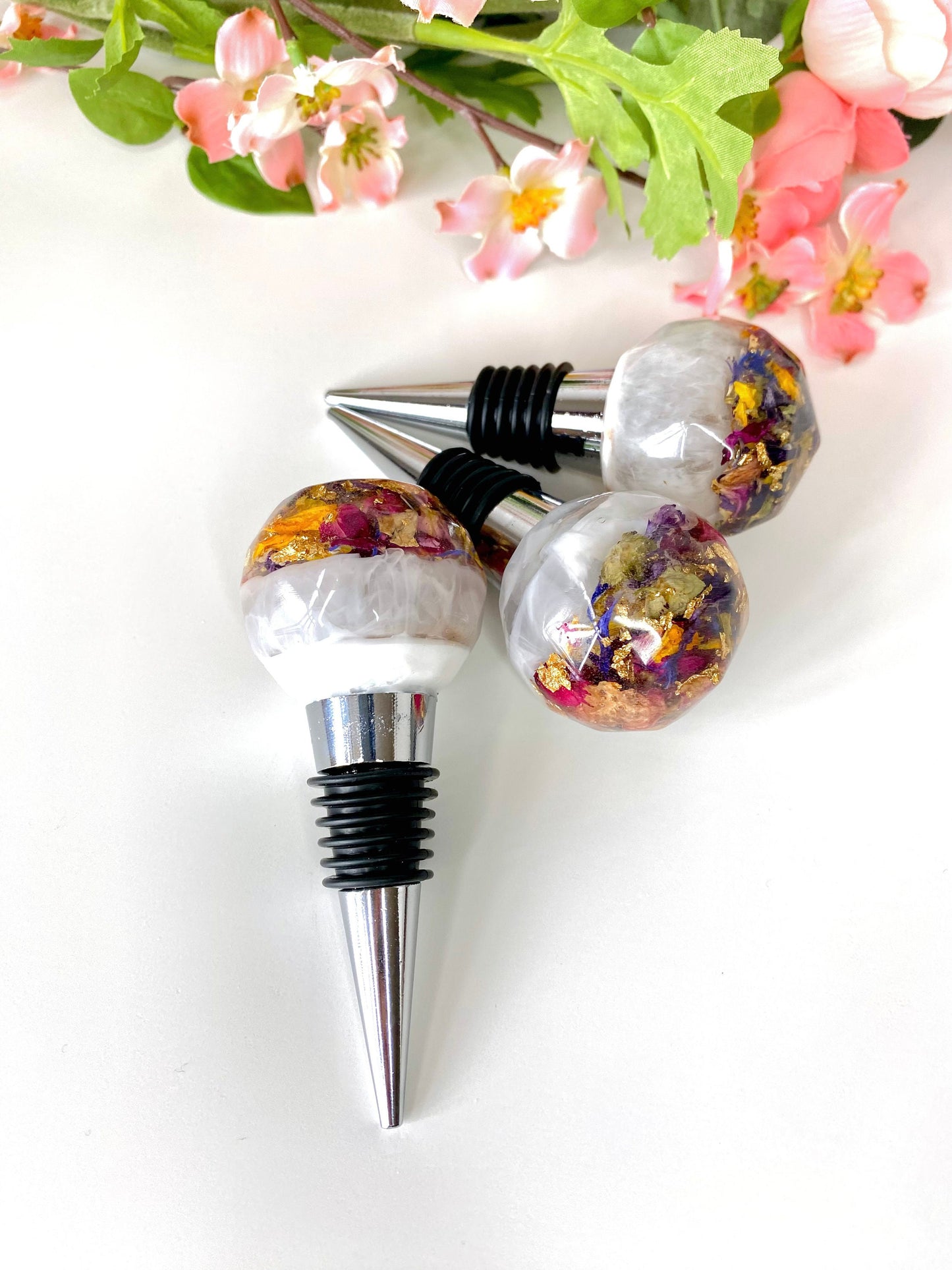 Close-up of handmade resin bottle stoppers with stainless steel bases, white resin tops with clear resin containing colourful dried flowers and gold leaf, highlighting intricate handmade details, decorative and functional wine accessories for gifts or home use