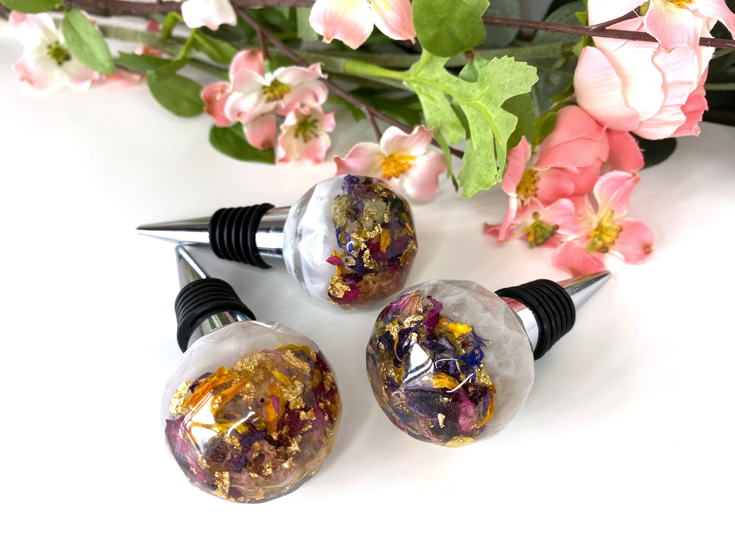 Close-up of three handmade resin bottle stoppers with stainless steel bases, white resin tops with clear resin containing colourful dried flowers and gold leaf, decorative and functional wine accessories for gifts or home use