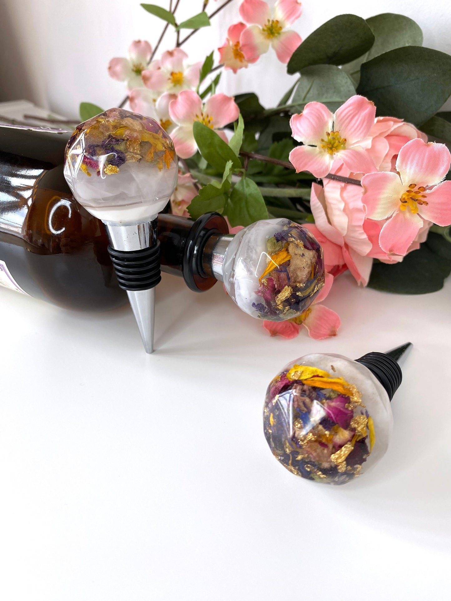 Three handmade resin bottle stoppers with stainless steel bases, white resin tops with clear resin containing colourful dried flowers and gold leaf, one inserted into a bottle lying flat on table, one balanced next to bottle showing full stopper, and one focusing on resin top, decorative wine accessories for gifts or home use.