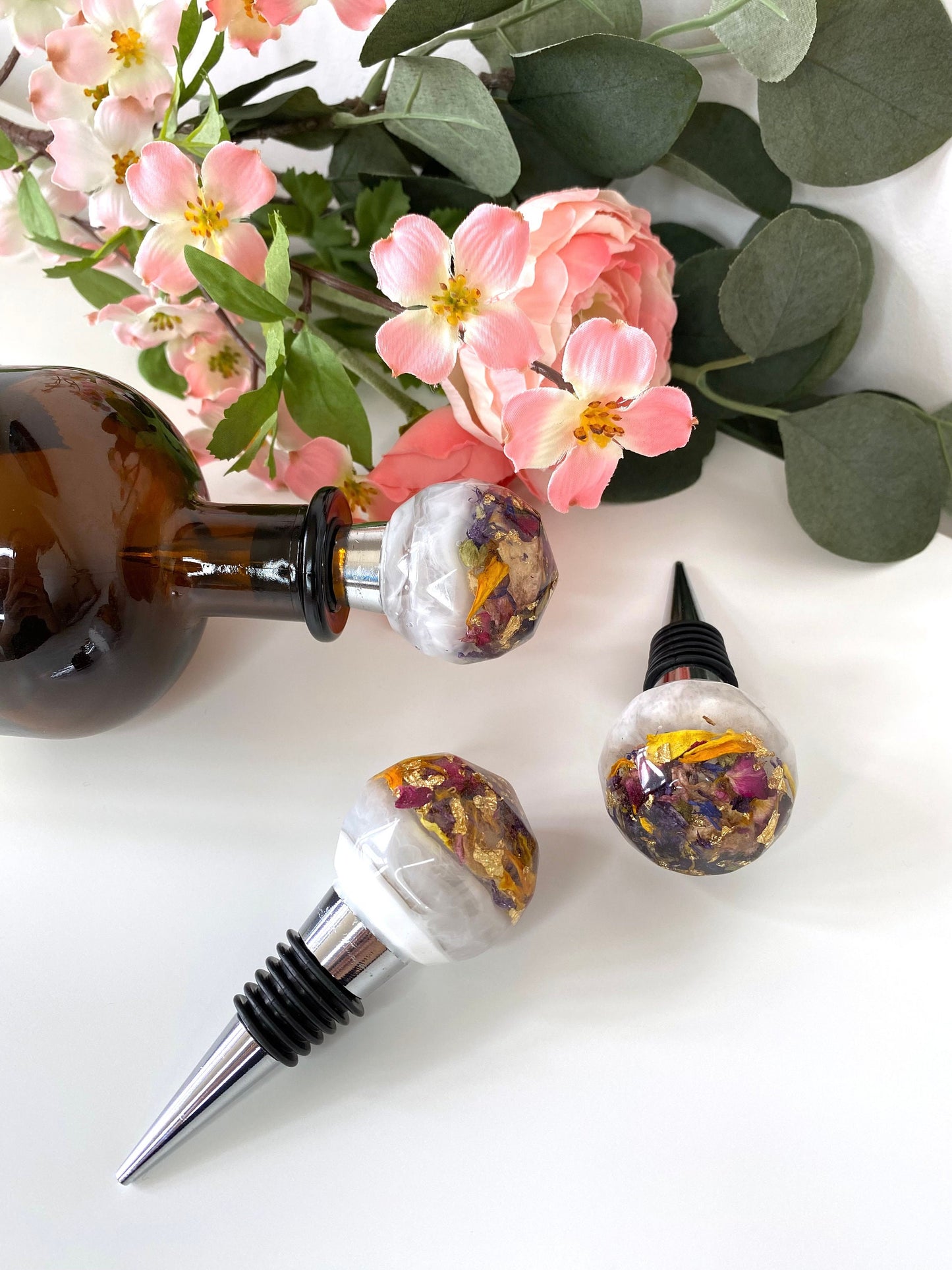 Three handmade resin bottle stoppers with stainless steel bases, white resin tops with clear resin containing colourful dried flowers and gold leaf, one inserted into a bottle lying flat on table, two lying flat next to the bottle showing full stoppers, decorative wine accessories for gifts or home use