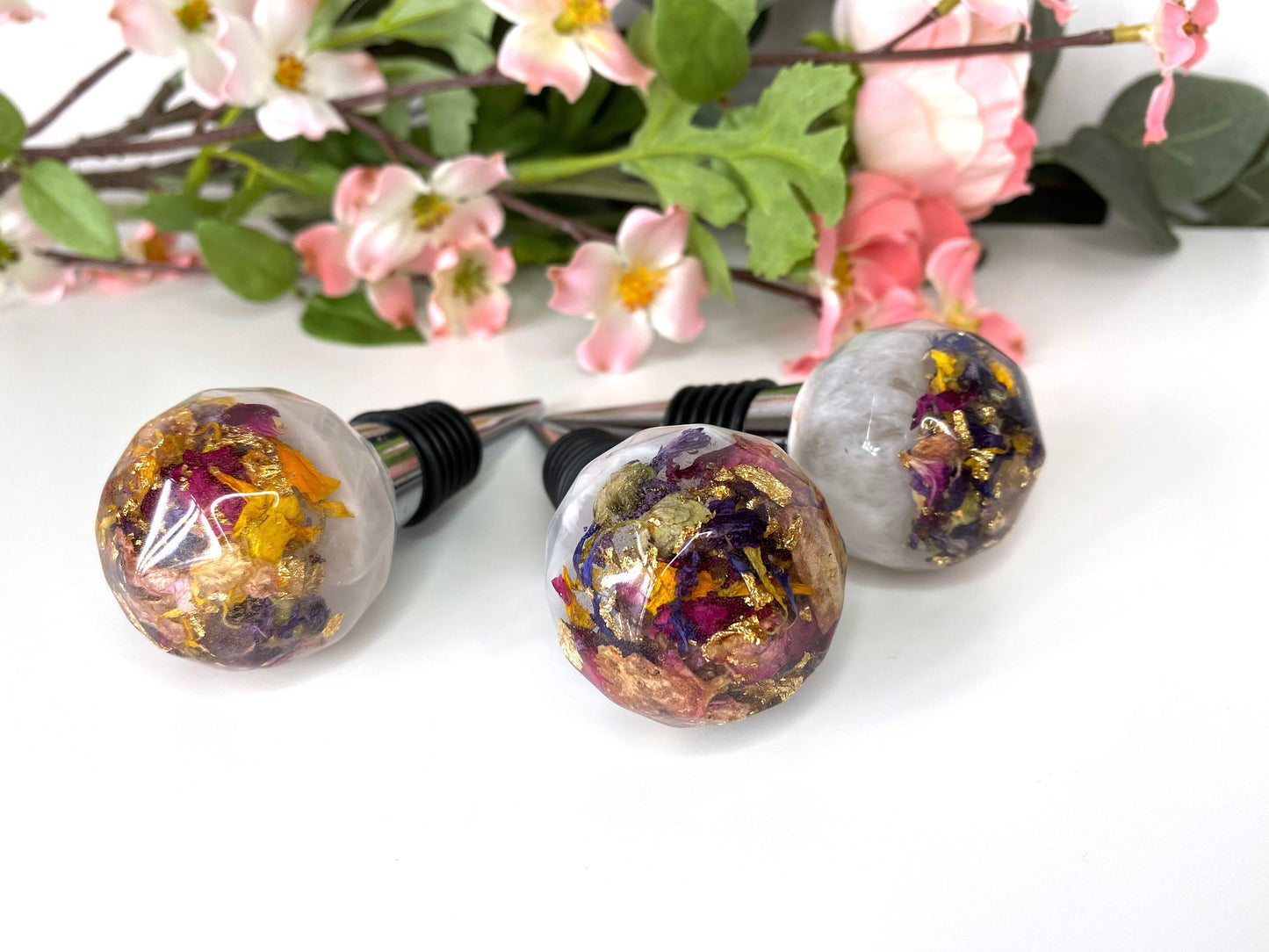 Three handmade resin bottle stoppers with stainless steel bases, white resin tops with clear resin containing colourful dried flowers and gold leaf, displayed in front of flowers, showing one full stopper and two focusing on resin tops, decorative and functional wine accessories for gifts or home use