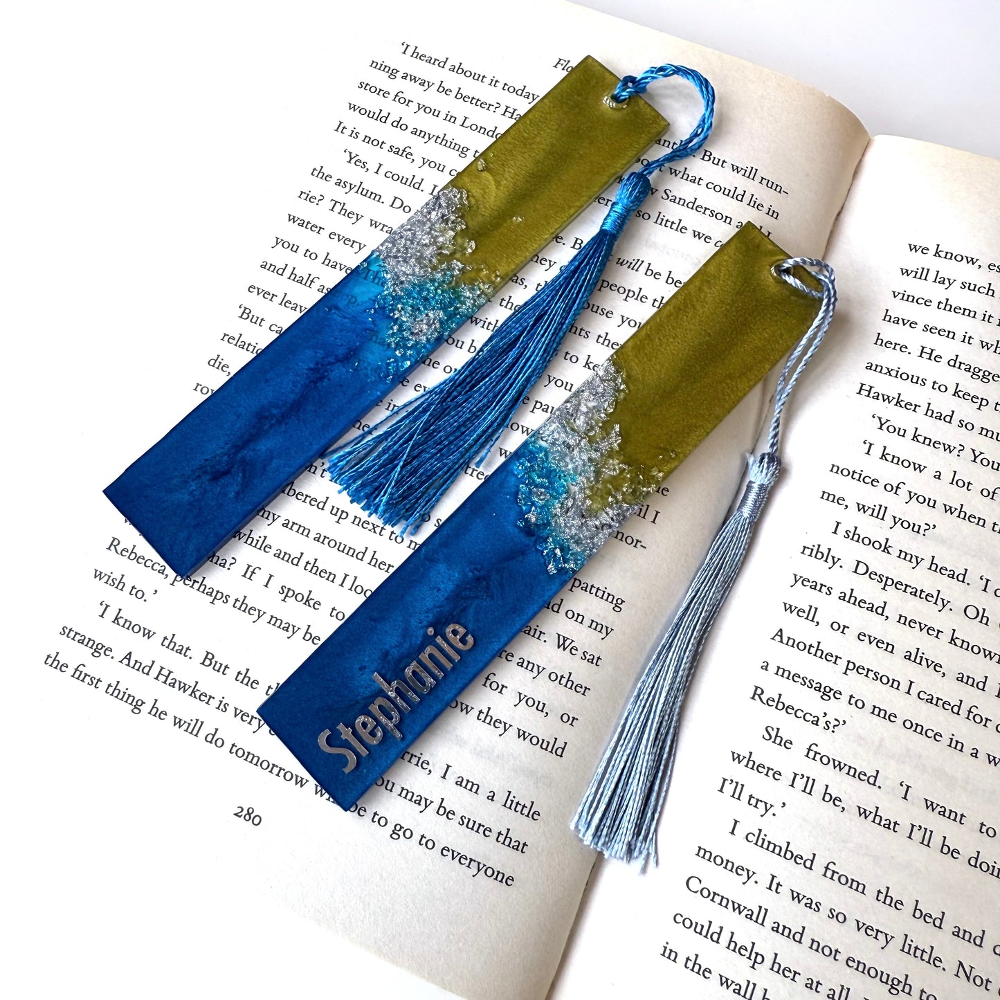 Two handmade resin bookmarks in an open book: one personalised with silver vinyl name, one plain. Blue, green, and silver celestial-inspired design, perfect gift for book lovers.