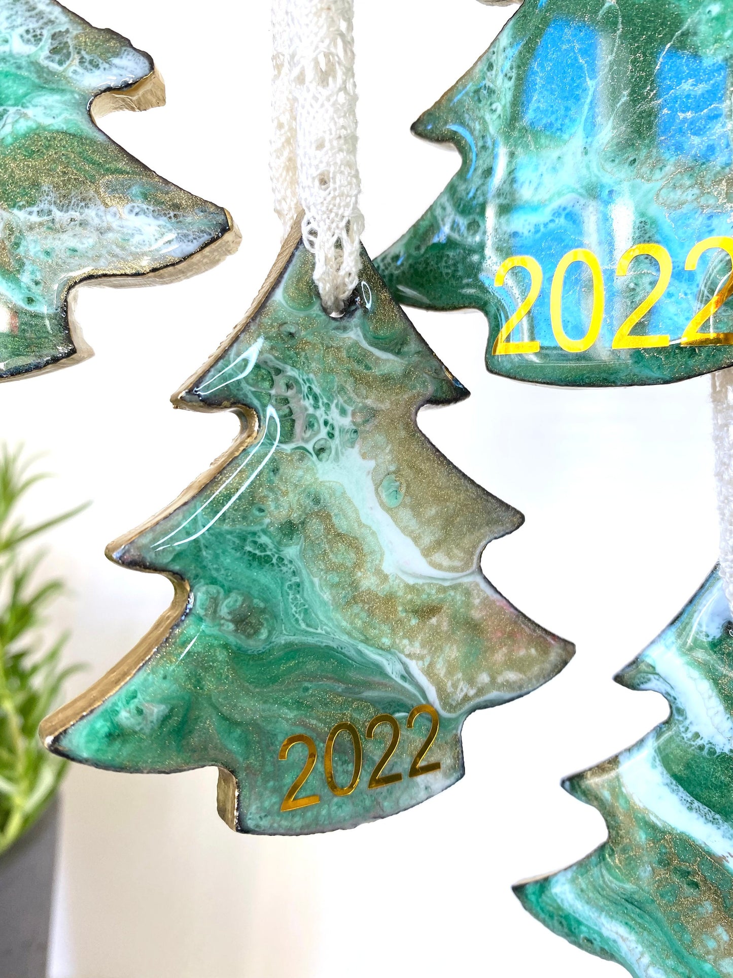 Close-up of a tree-shaped resin Christmas ornament on slate, green, white, and bronze/gold resin design, gold-painted edges, beige lace ribbon, personalised with 2025, with other ornaments blurred in the background.