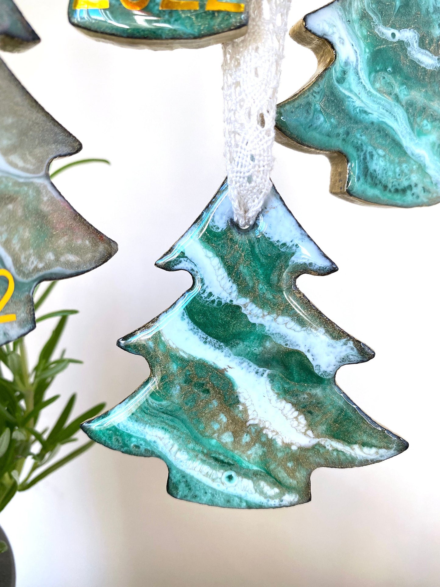 Close-up of a tree-shaped resin Christmas ornament on slate, featuring green, white, and bronze/gold resin design with gold-painted edges and beige lace ribbon, non-personalised, other ornaments softly blurred in background.