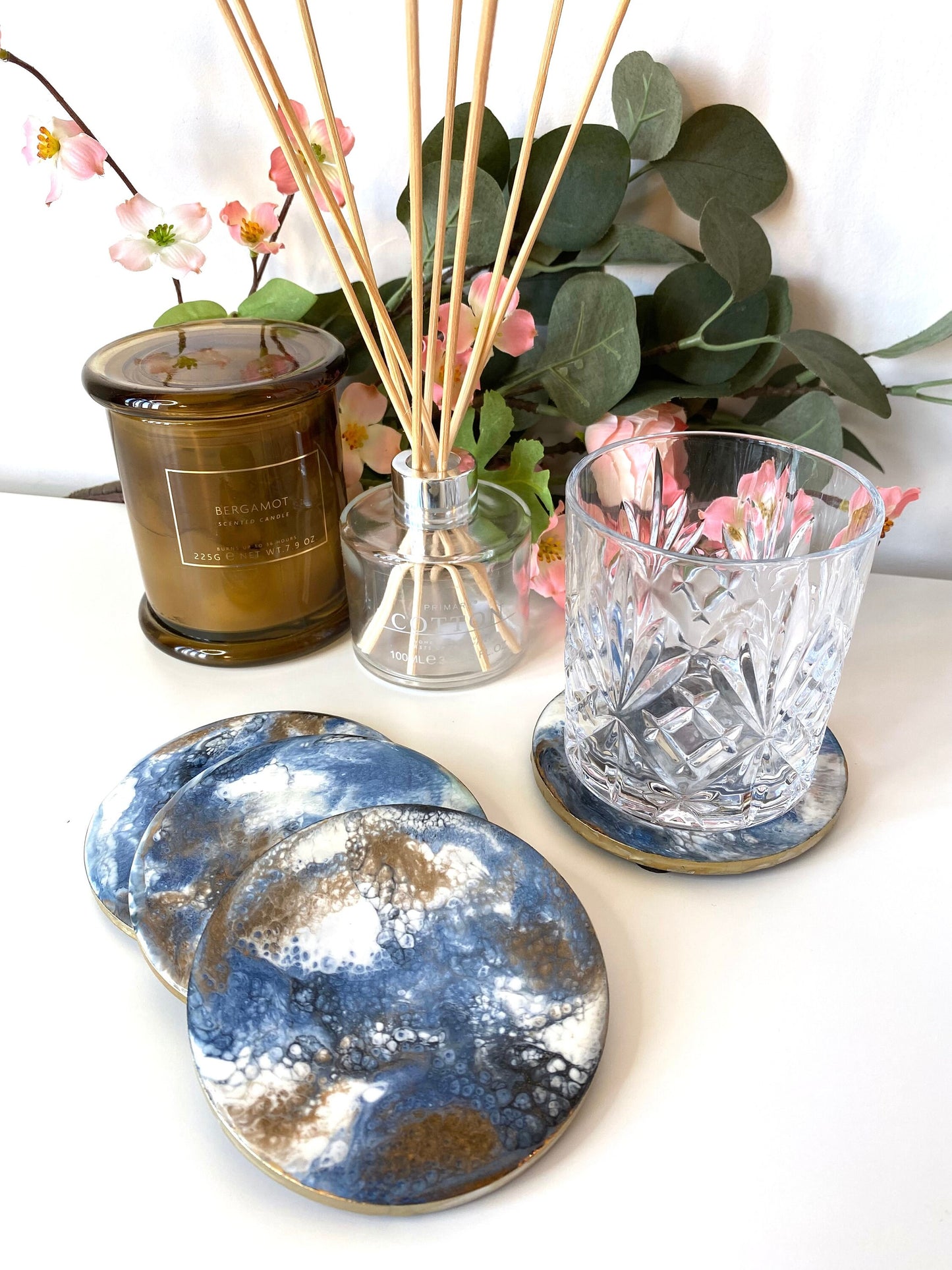 Set of 4 handmade round slate resin coasters with blue, white, black, and gold epoxy design and gold-painted edges, three stacked domino-style with the fourth holding a water glass – elegant home décor
