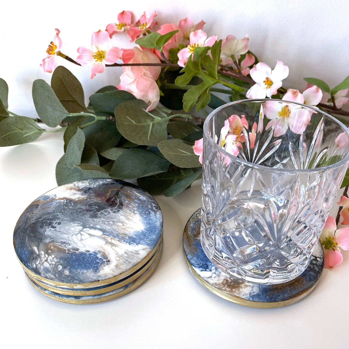 Set of 4 handmade round slate resin coasters with blue, white, black, and gold epoxy design and gold-painted edges, three stacked with the fourth next to them holding a water glass