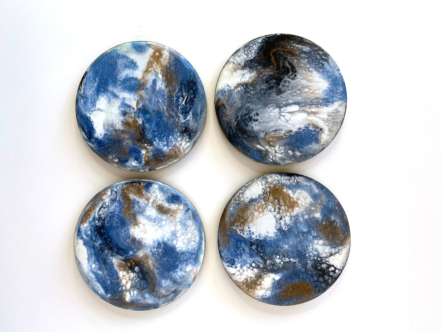 Top-down view of 4 handmade round slate resin coasters with blue, white, black, and gold epoxy designs and gold-painted edges – each coaster is unique and one-of-a-kind.